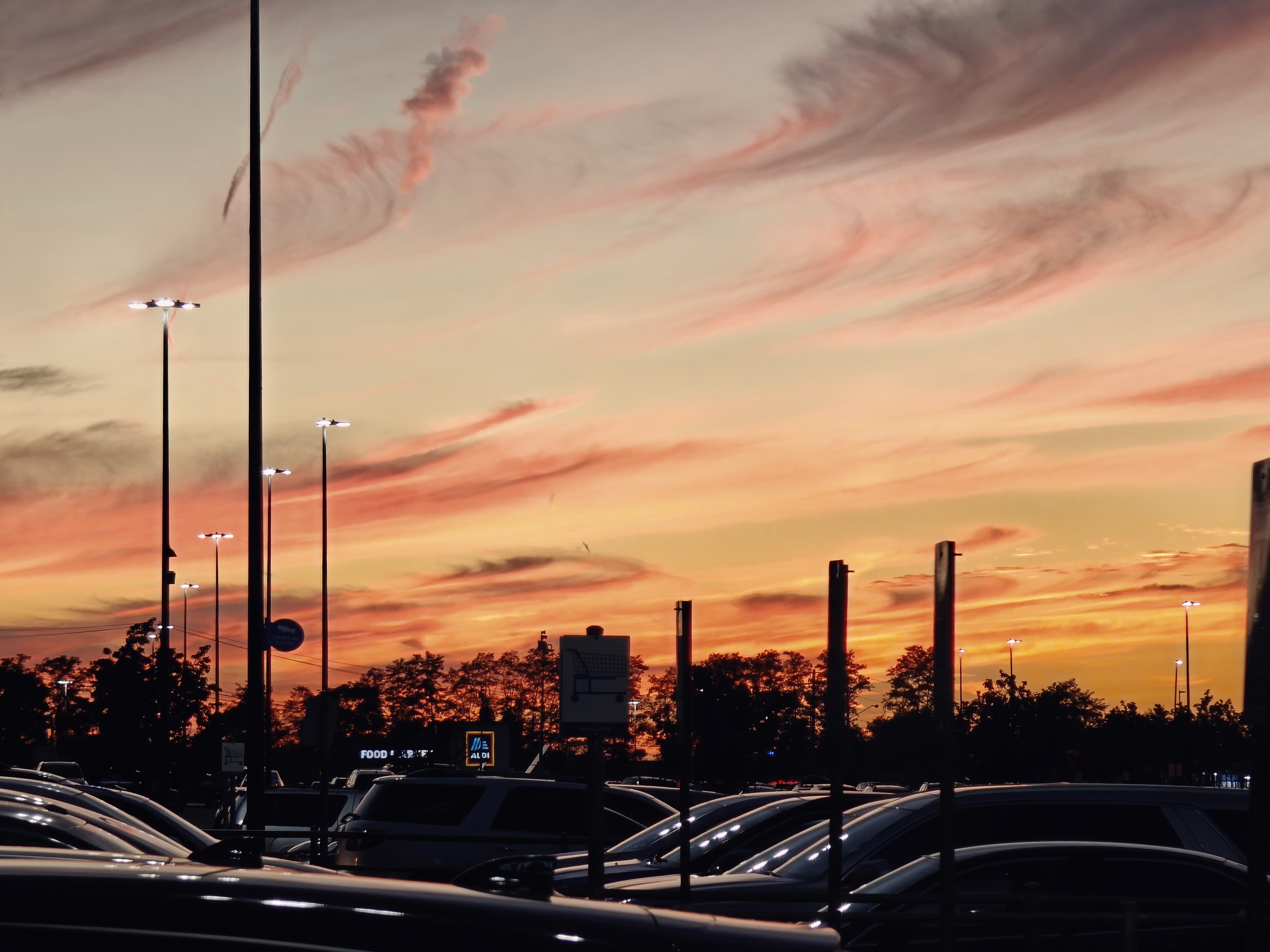 An orange-pink sky silhouettes repeating parking-lot lamps, cars, and a low suburban horizon.