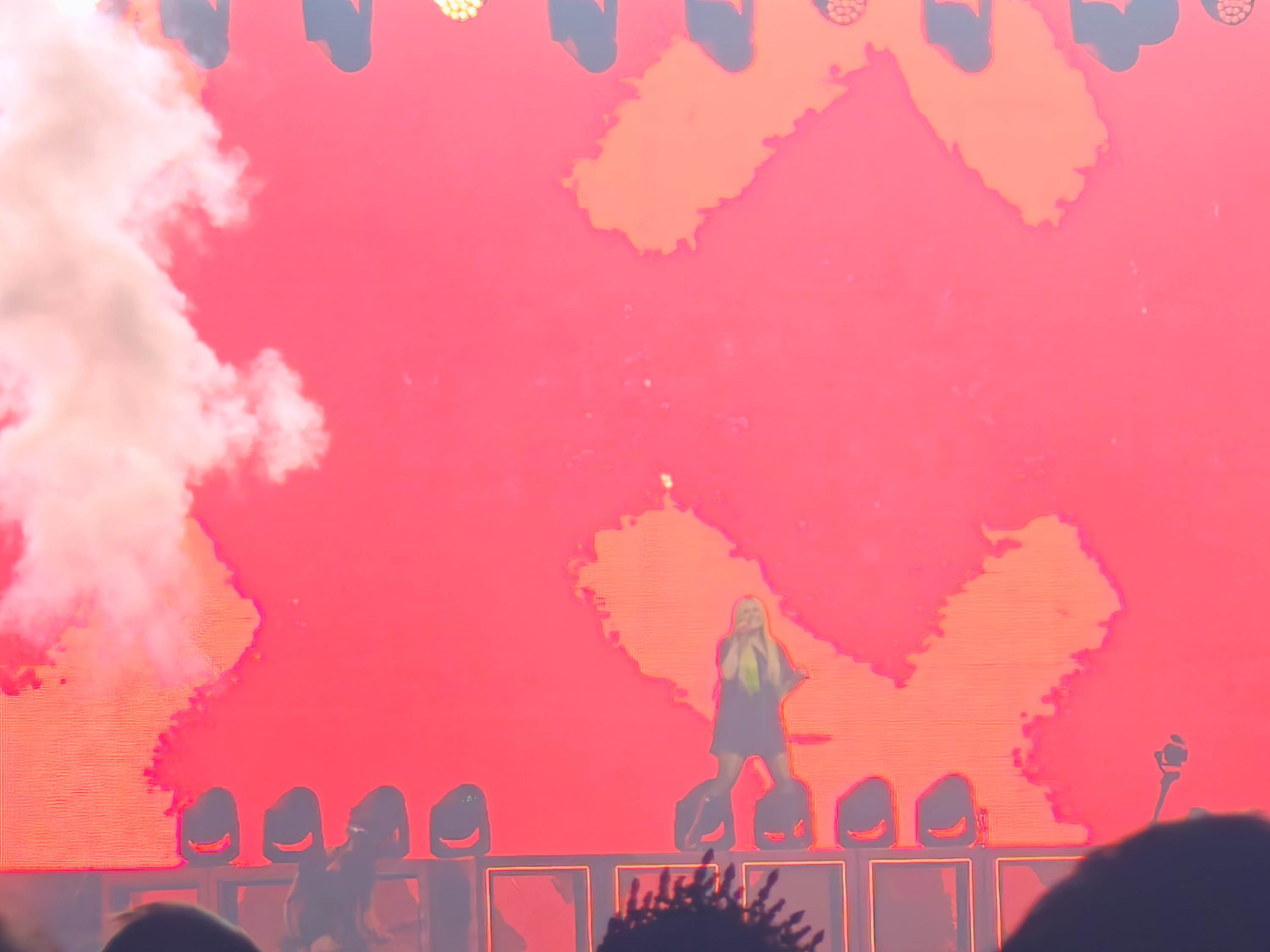 A singer appears small against an immense hot-pink stage screen while white smoke rises at left.
