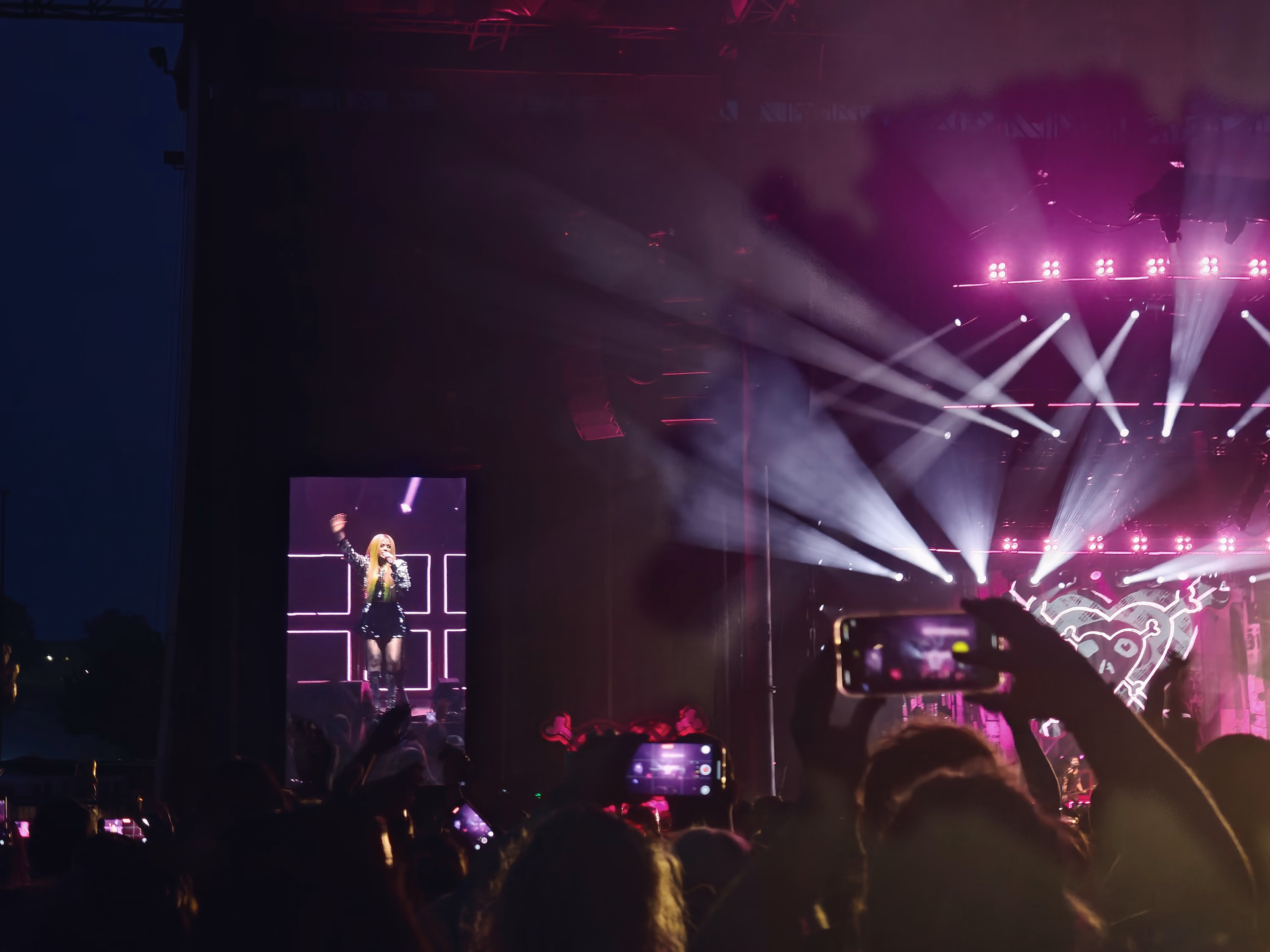 Magenta and white stage beams cross above raised phones and a concert crowd at Hersheypark Stadium.