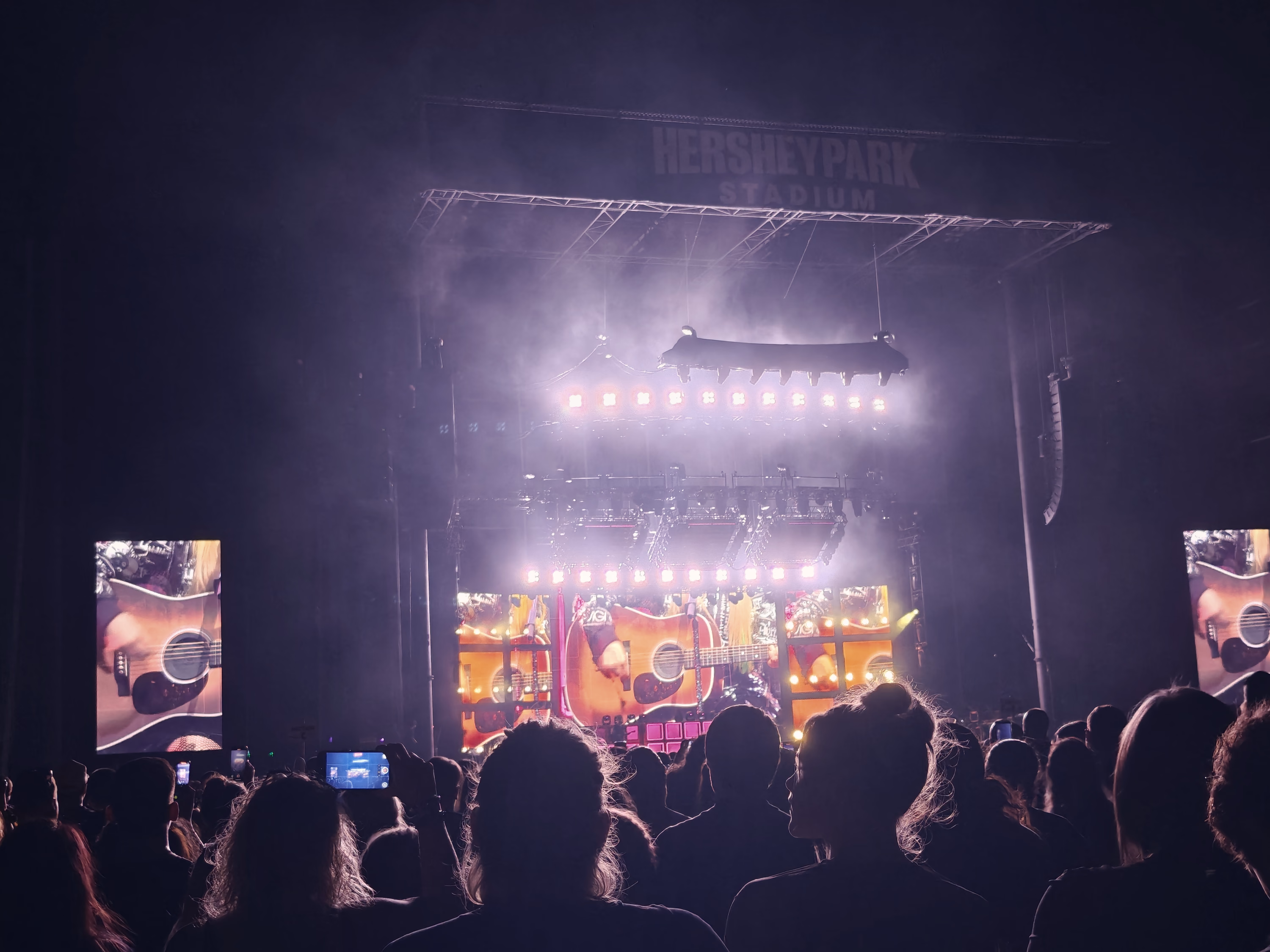 A bright guitar fills the stadium screens while violet haze hangs above a dark field of audience silhouettes.