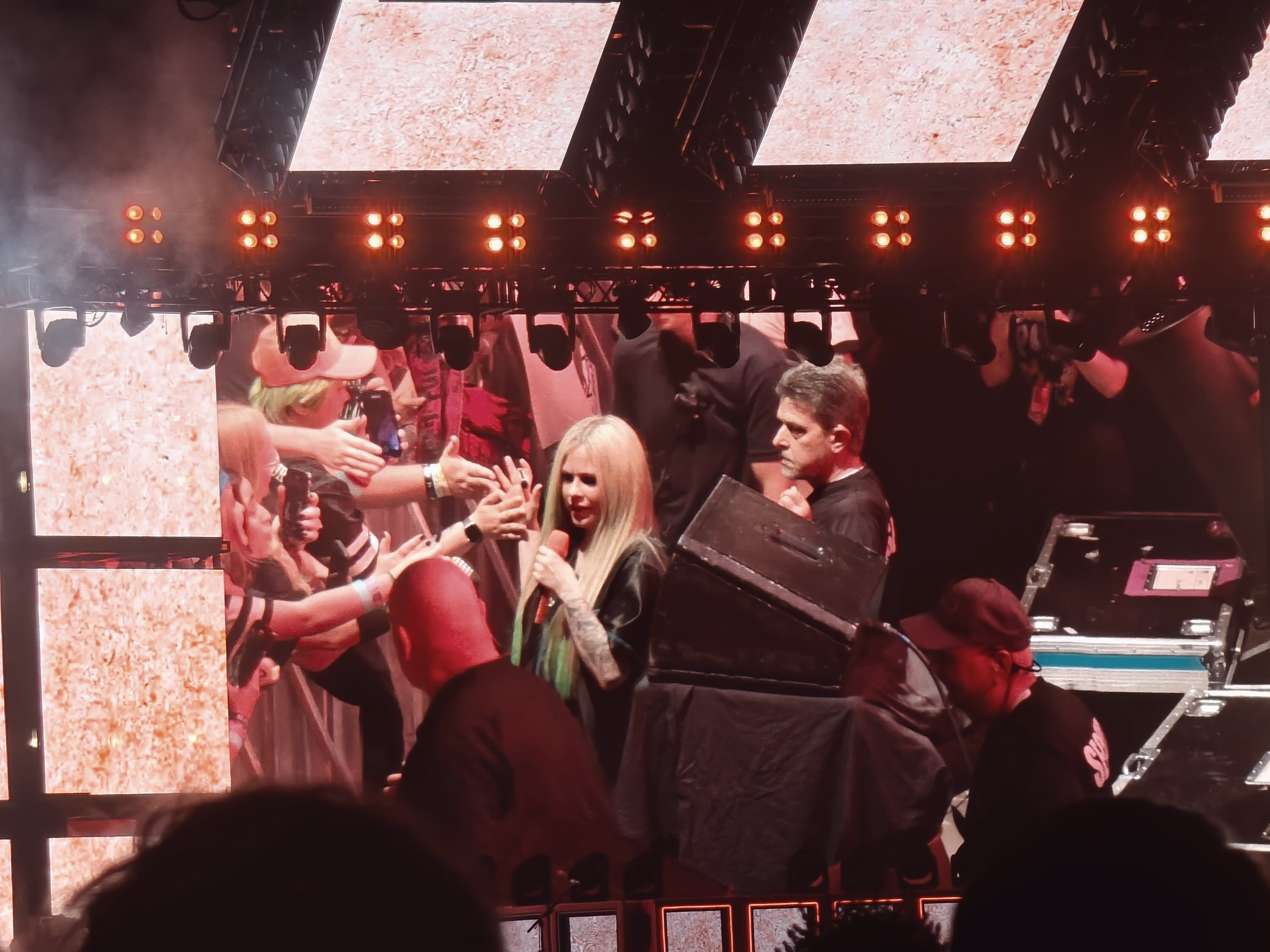 A singer reaches toward fans at the barricade under red stage light while crew and equipment crowd the scene.