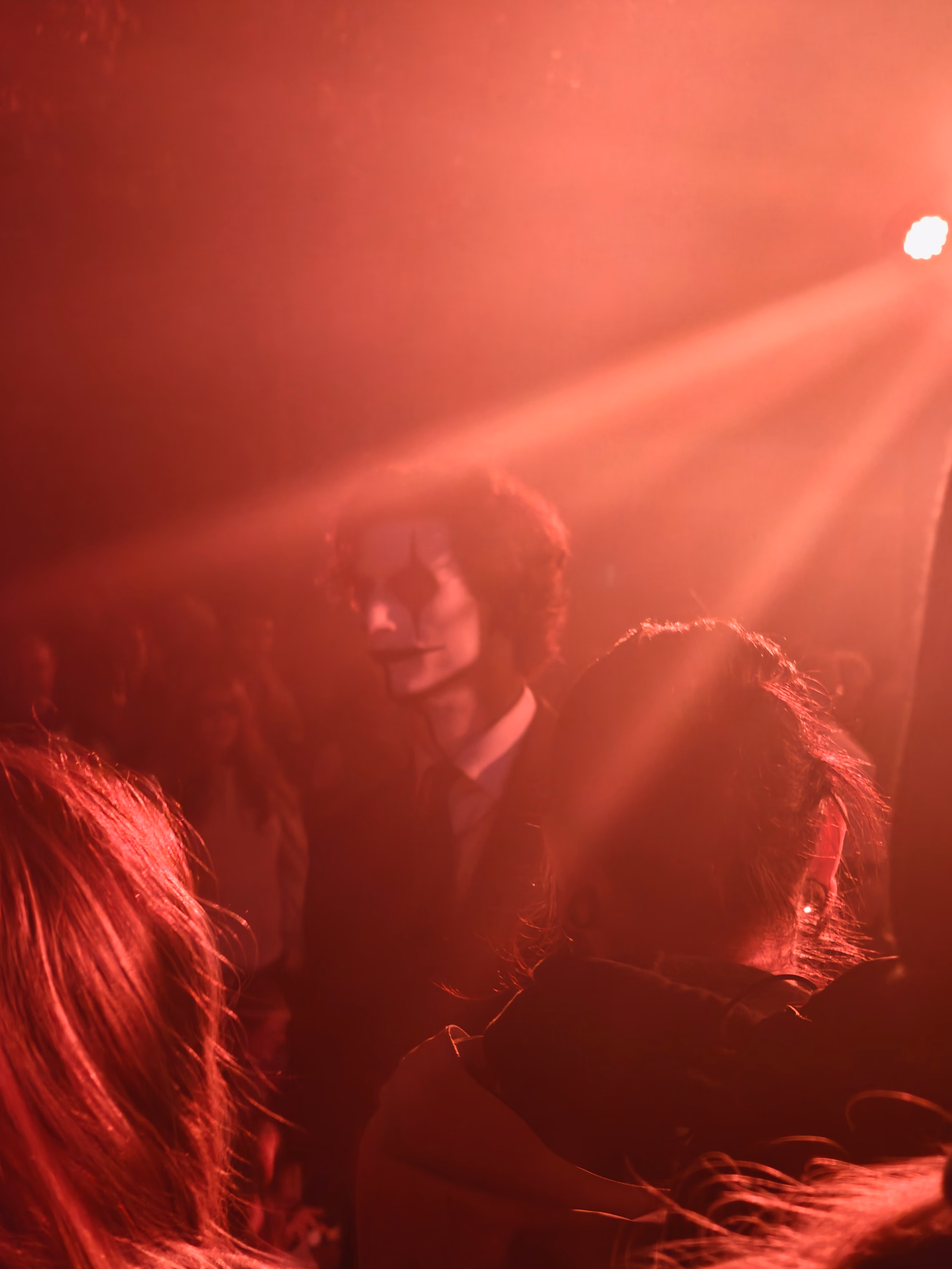 A face emerges from a red fog-filled crowd beneath a single diagonal spotlight.