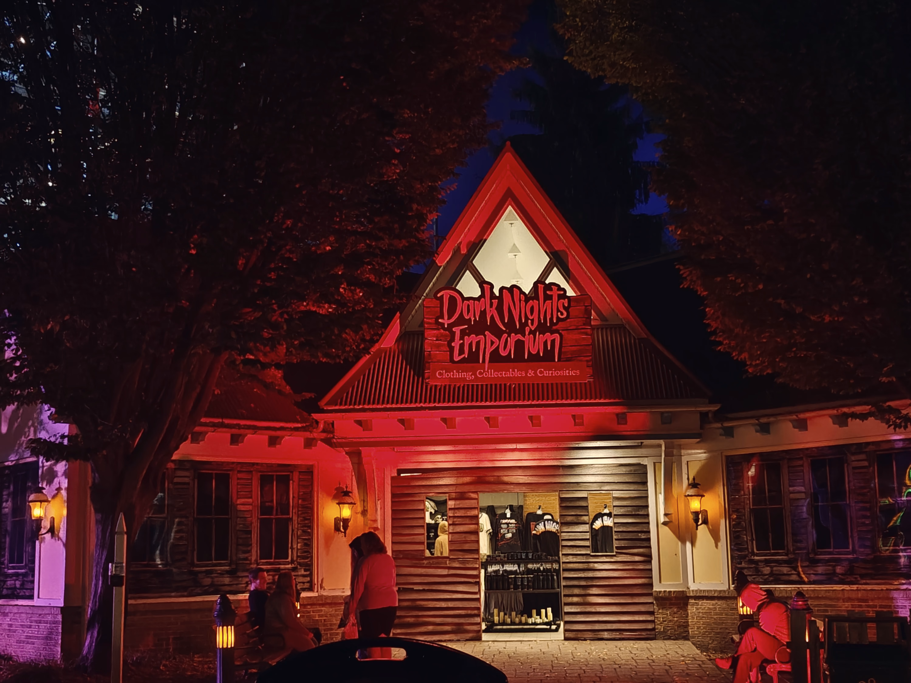 A small peaked storefront glows red beneath trees around the Dark Nights Emporium sign.