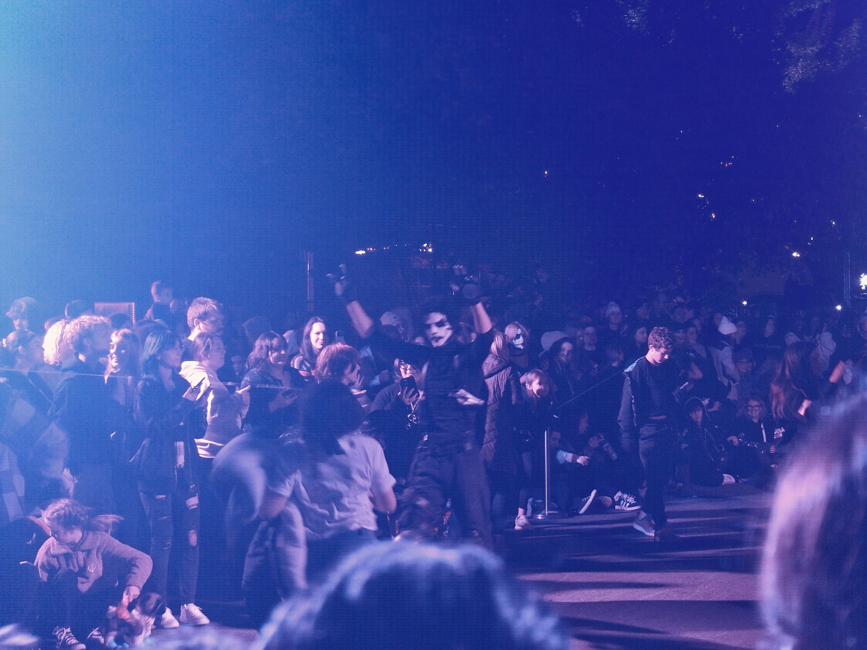 A moving performer lifts a prop above a blue-lit crowd while haze and foreground figures blur the edges.