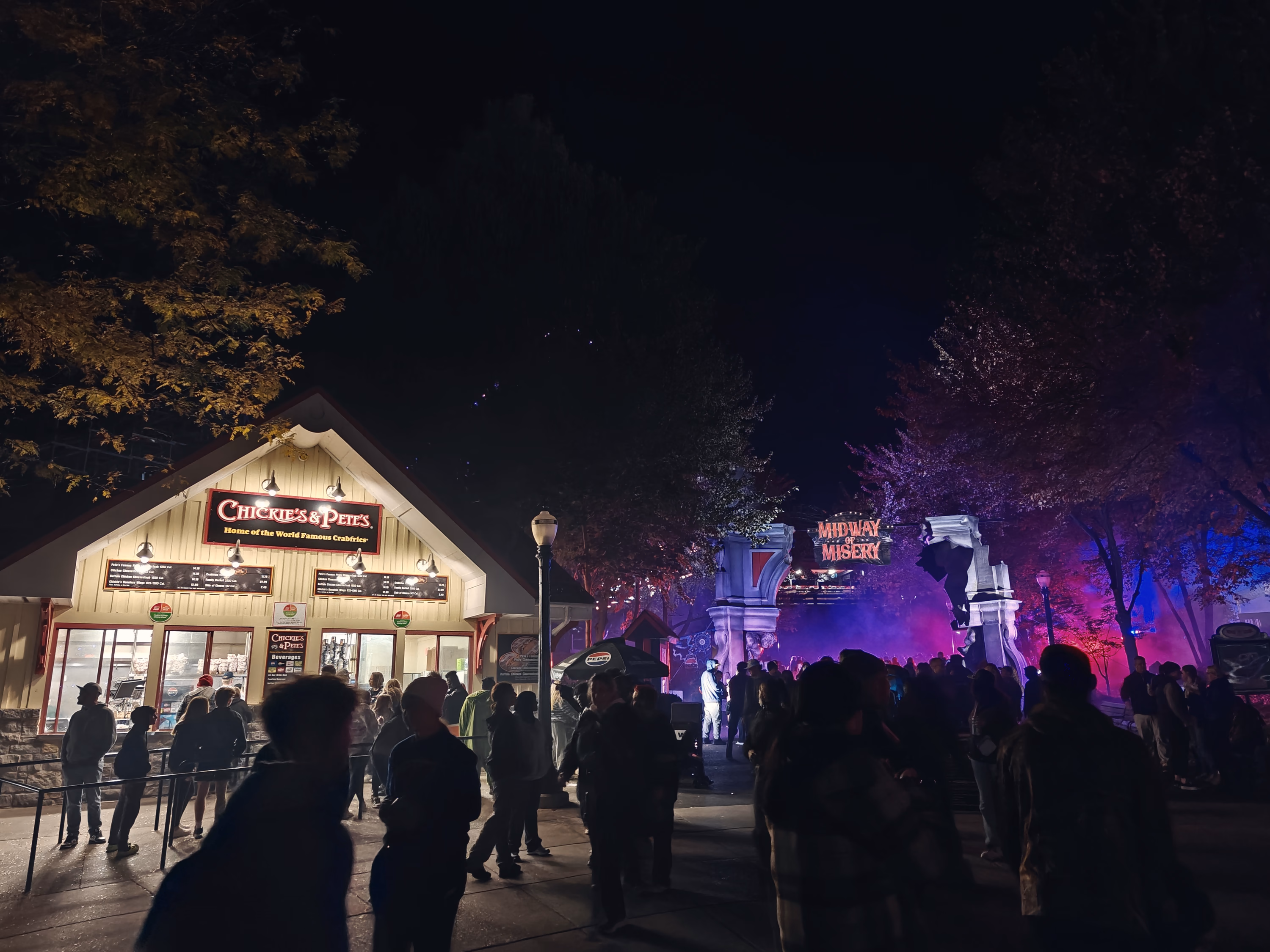 A food stand and the distant Midway of Misery split a crowded night street into warm white and violet-red light.