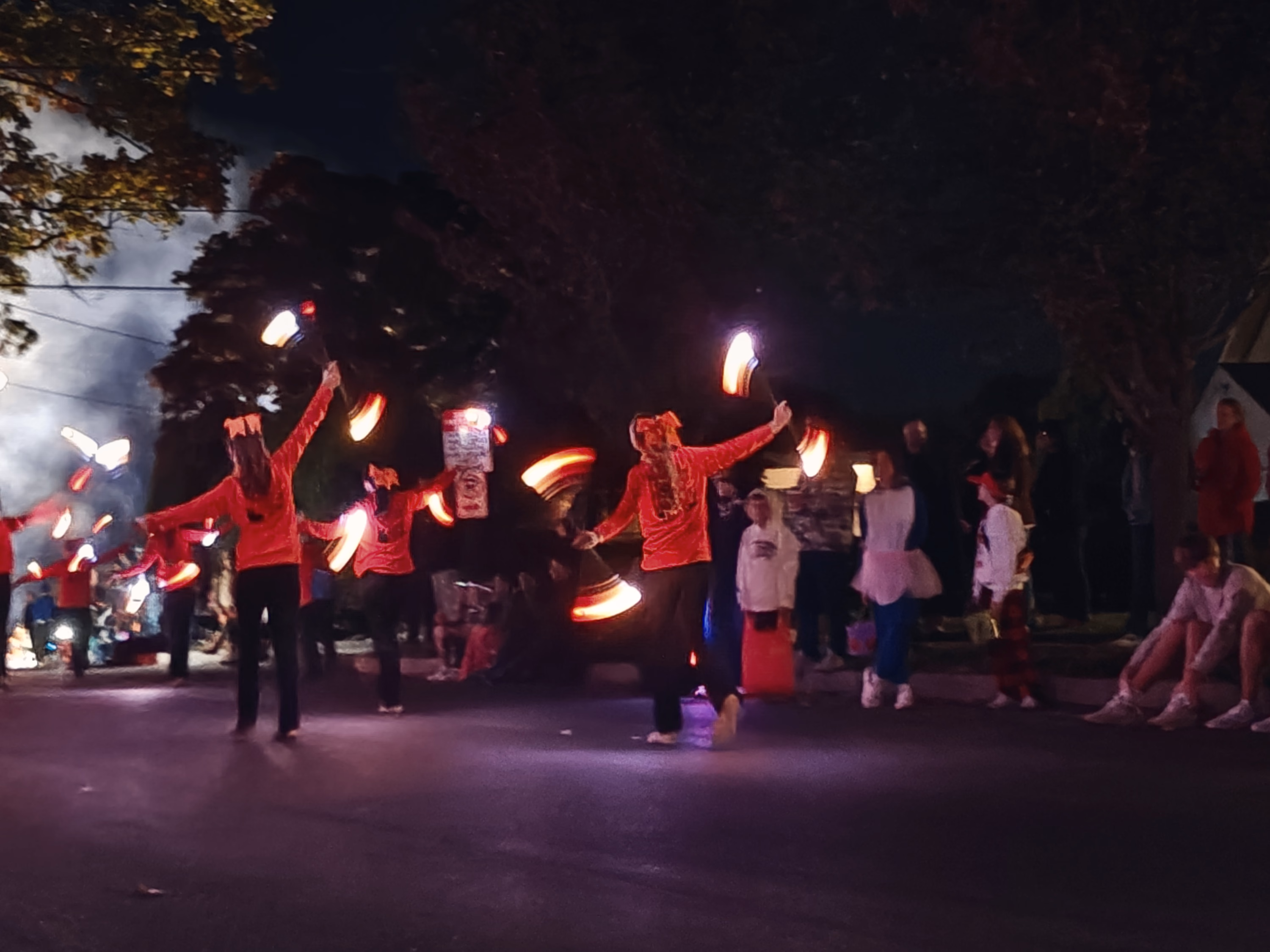 Parade dancers in red swing glowing batons along a residential street lined with seated spectators.