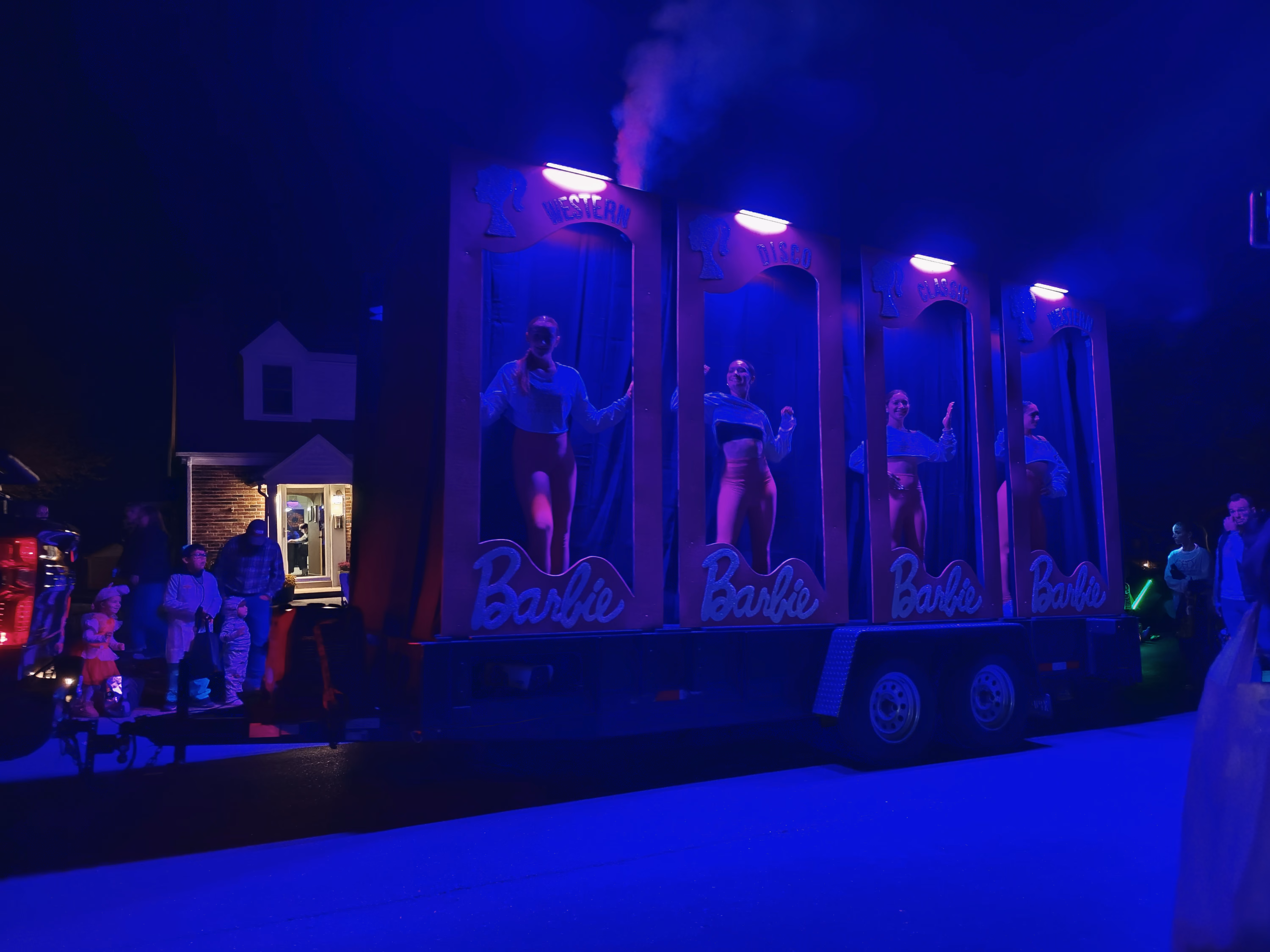Four performers stand in tall Barbie boxes on a blue and violet parade float as families watch from the curb.