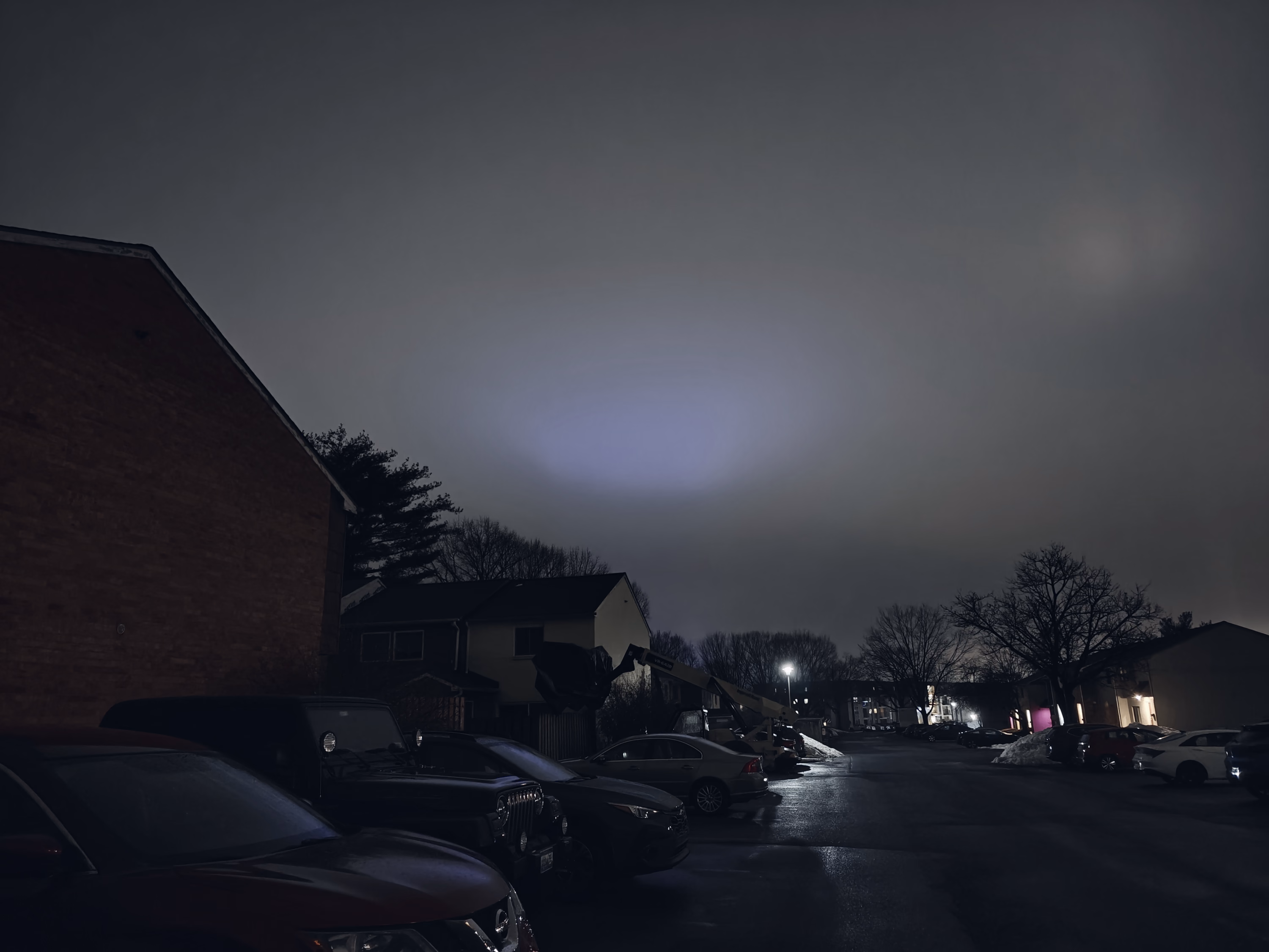A wet residential parking lot, cars, bare trees, and snowbanks sit beneath a low gray sky with an unexplained blue glow.