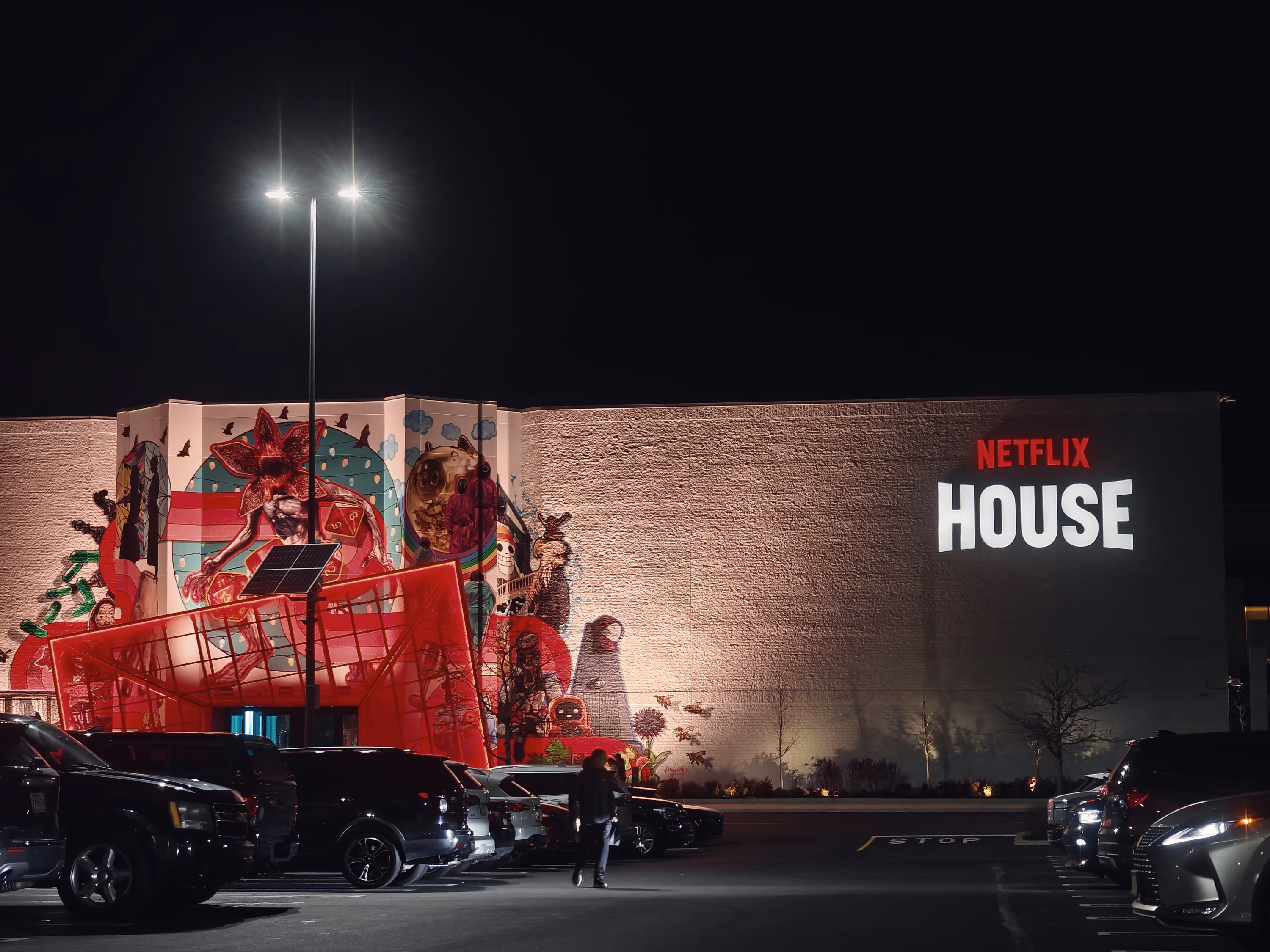 A lone pedestrian crosses between rows of cars before a red-lit mural and Netflix House sign at night.
