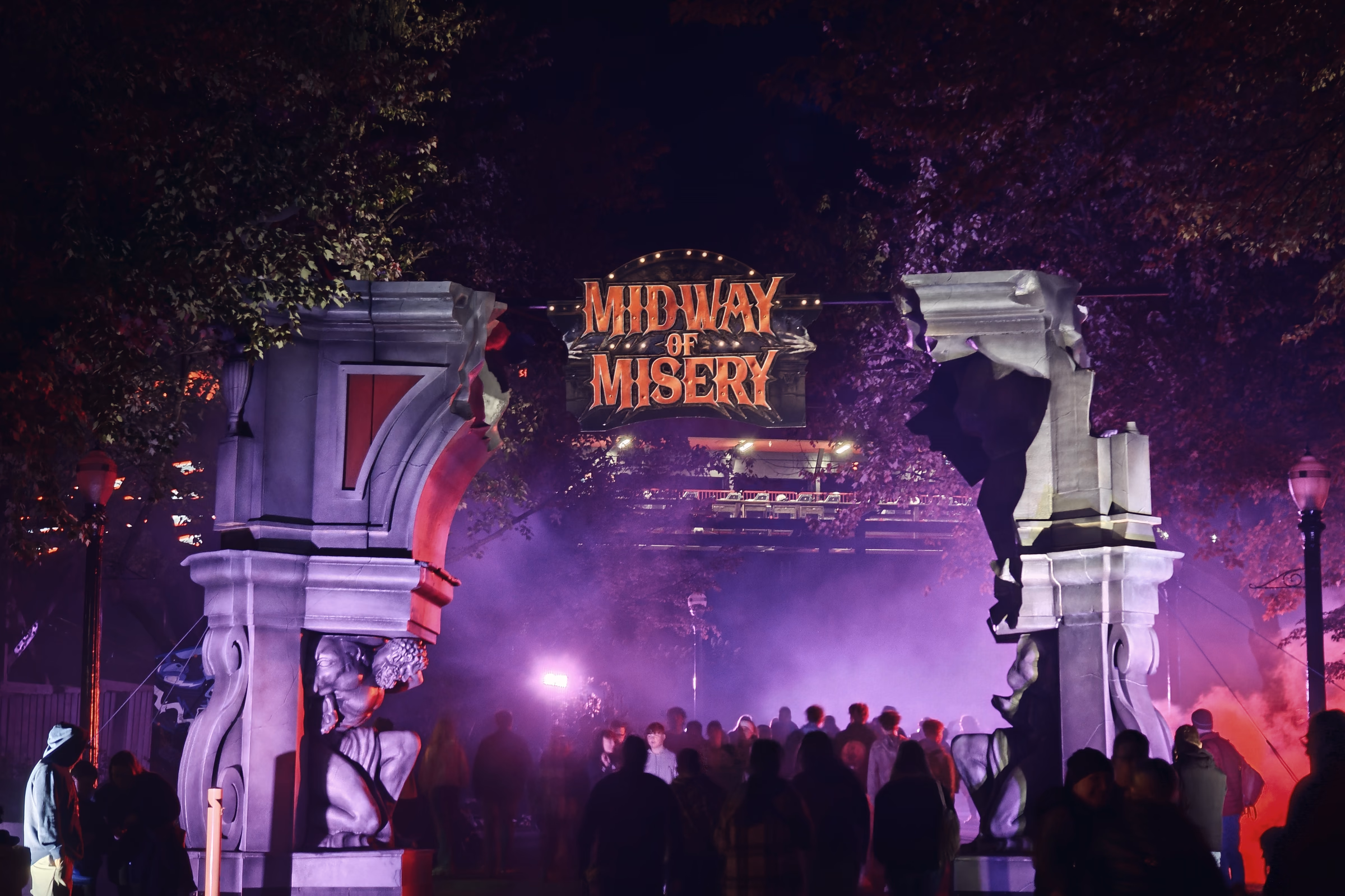 A broken-stone arch opens onto a crowd and dense violet fog beneath the Midway of Misery sign.