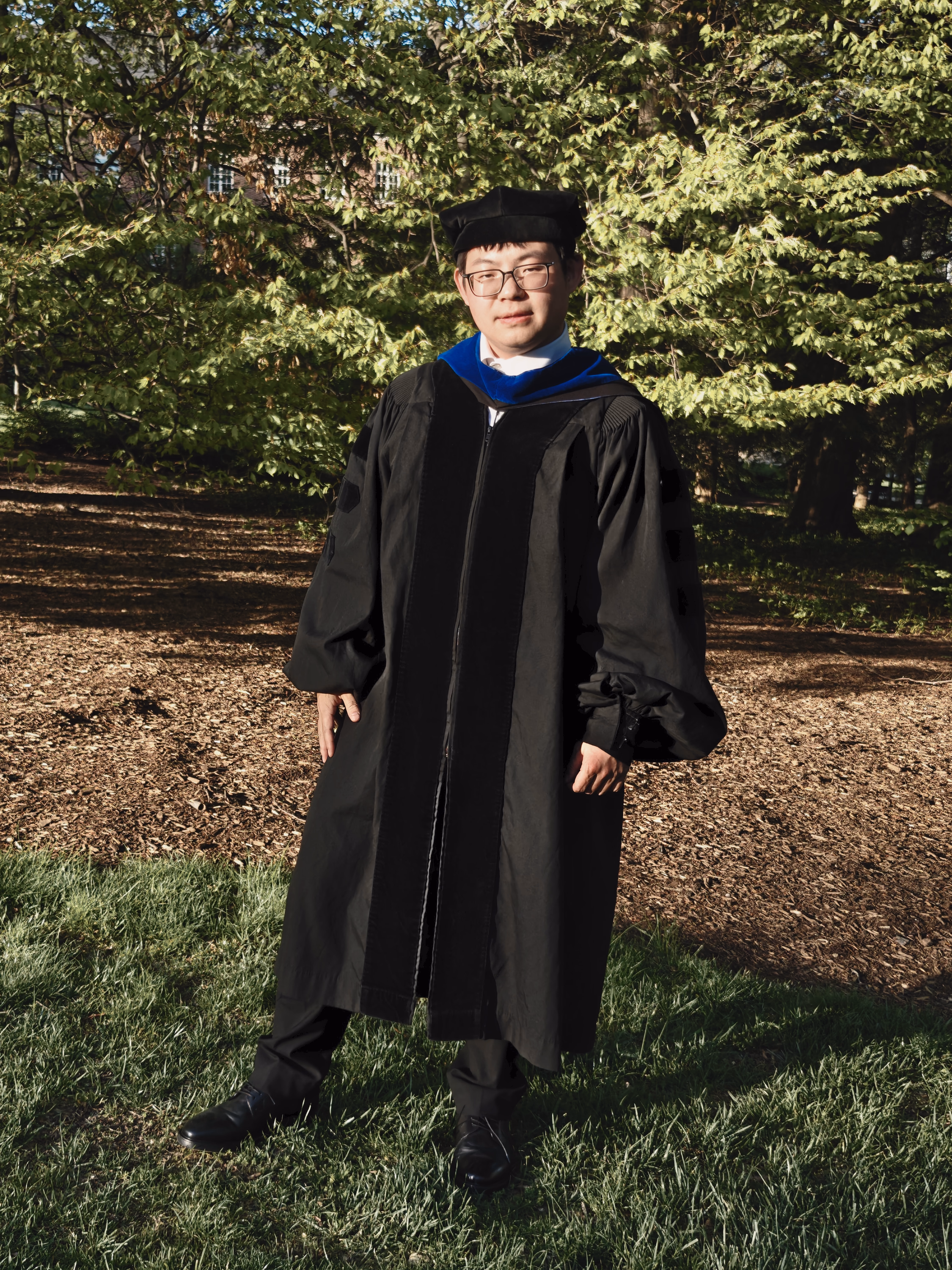 Full-length graduate in cap and gown standing on a sunlit campus hillside.