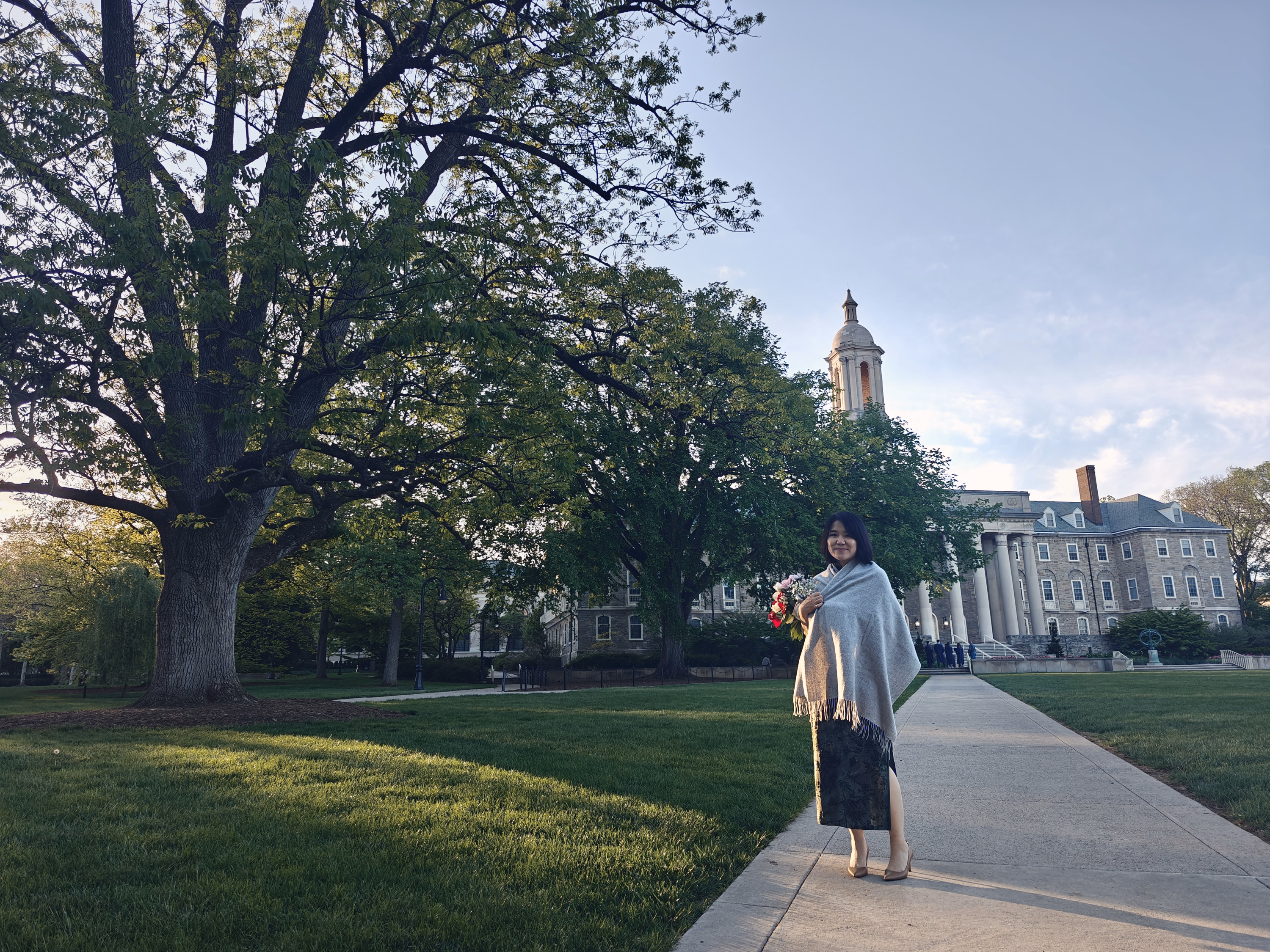 Woman holding flowers beneath a large tree with Penn State Old Main visible across the lawn.