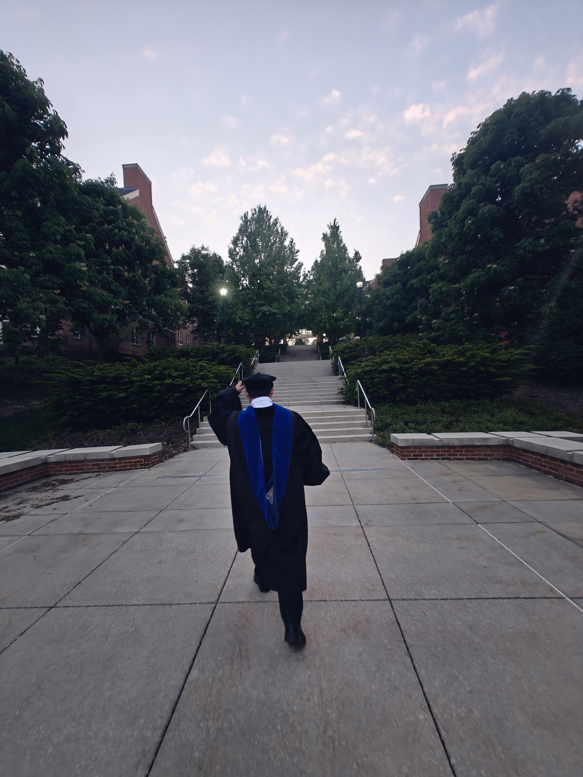 Graduate seen from behind walking toward broad campus stairs at dusk.
