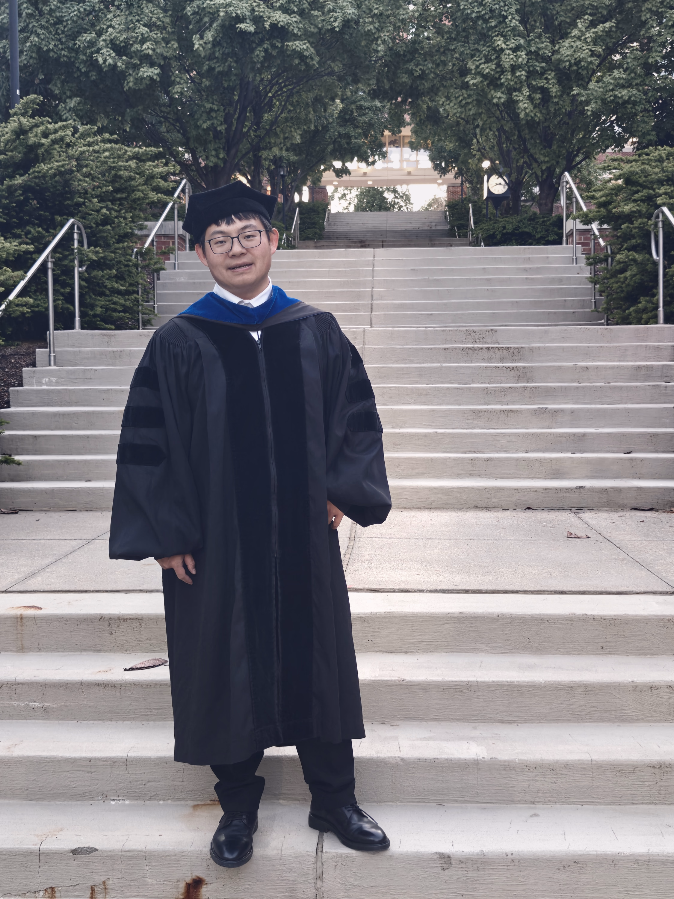 Full-length graduate facing the camera at the foot of broad Penn State campus stairs.