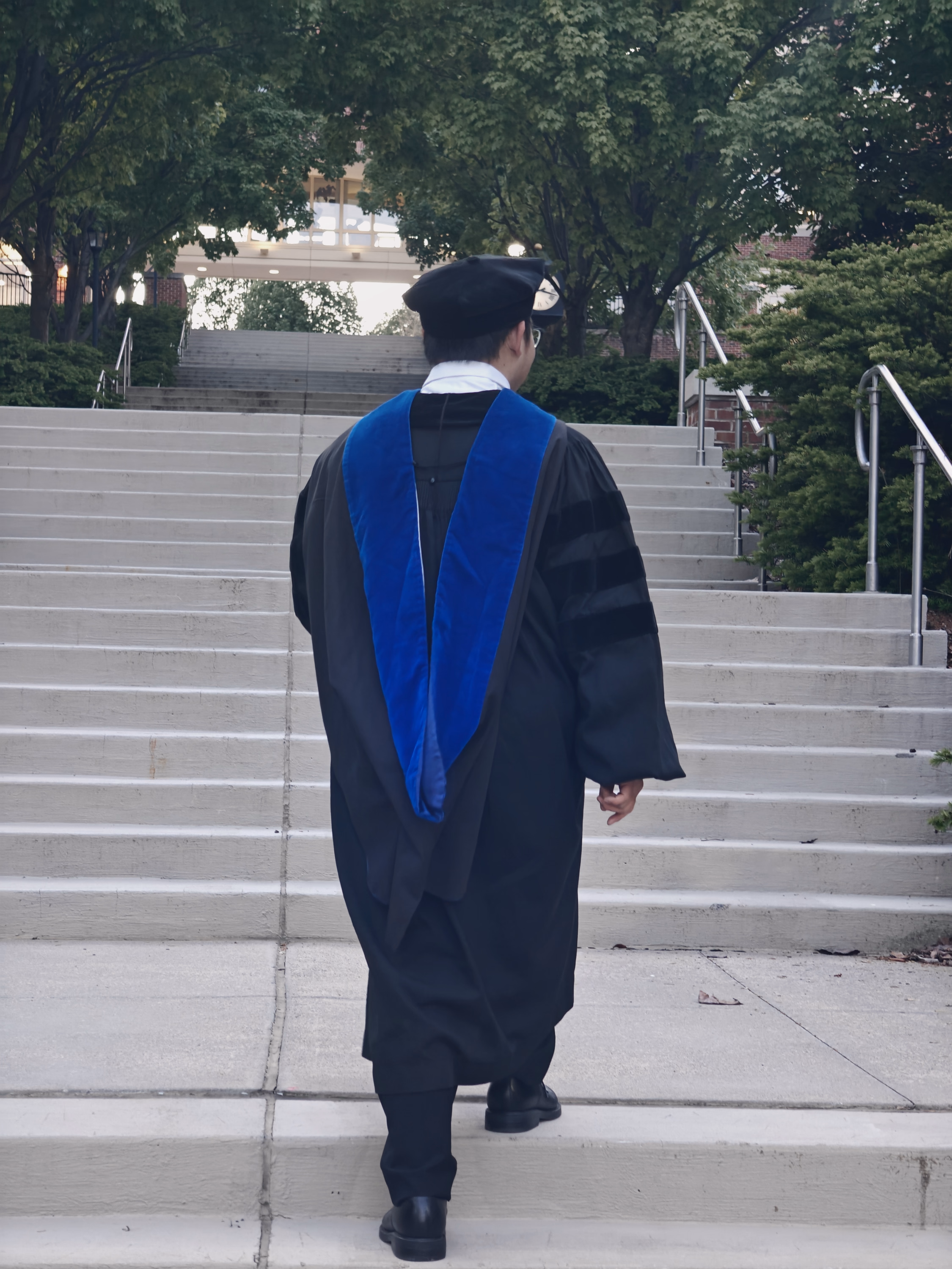 Back view of a graduate in black gown and bright blue hood walking up broad campus stairs.