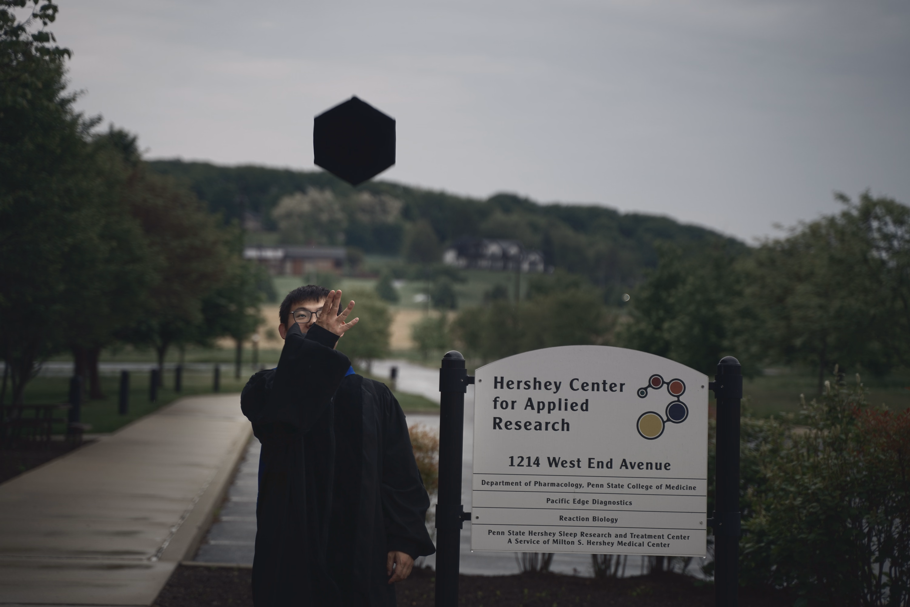 Graduate reaching toward a mortarboard suspended in the gray sky above the research center sign.
