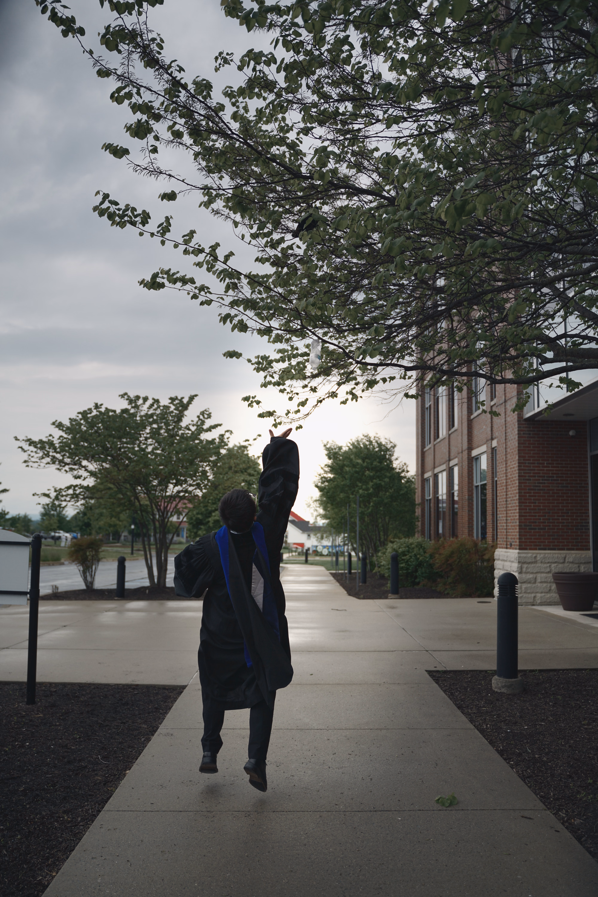 Graduate jumping and throwing a cap above a straight campus path under overcast light.