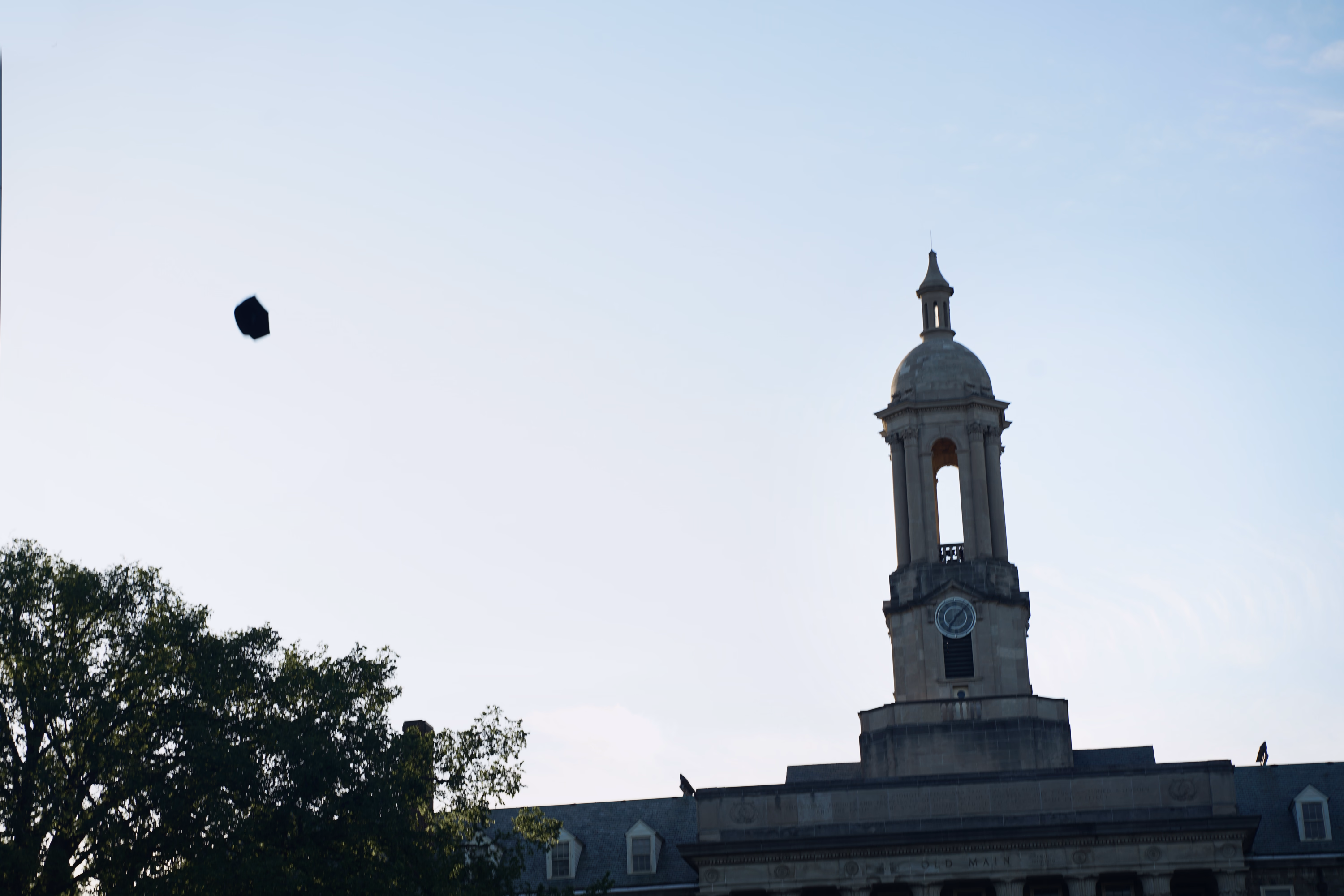 Mortarboard flying through a vivid blue sky above Old Main tower and a flag.