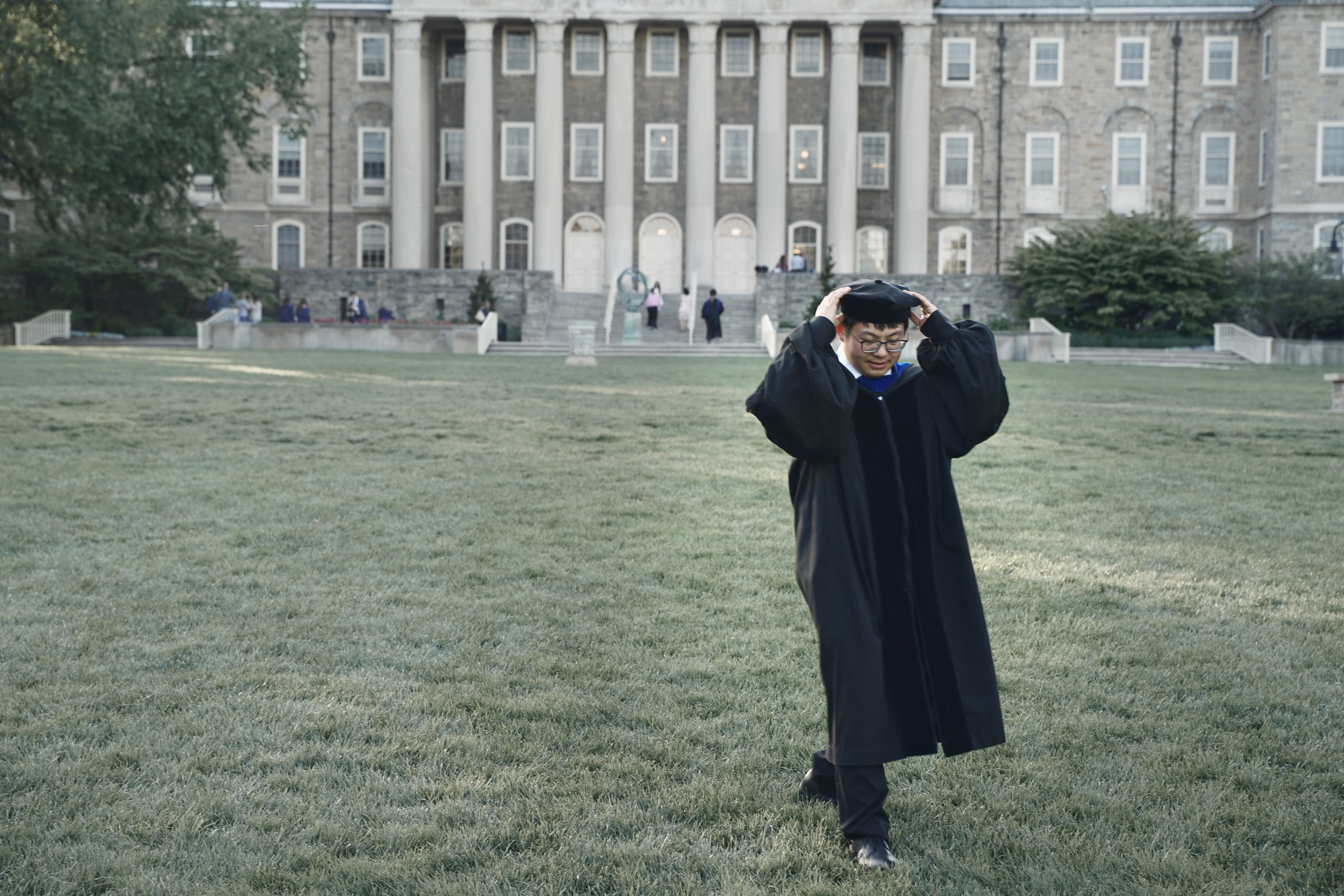 Graduate adjusting a mortarboard while walking across the lawn in front of Old Main.
