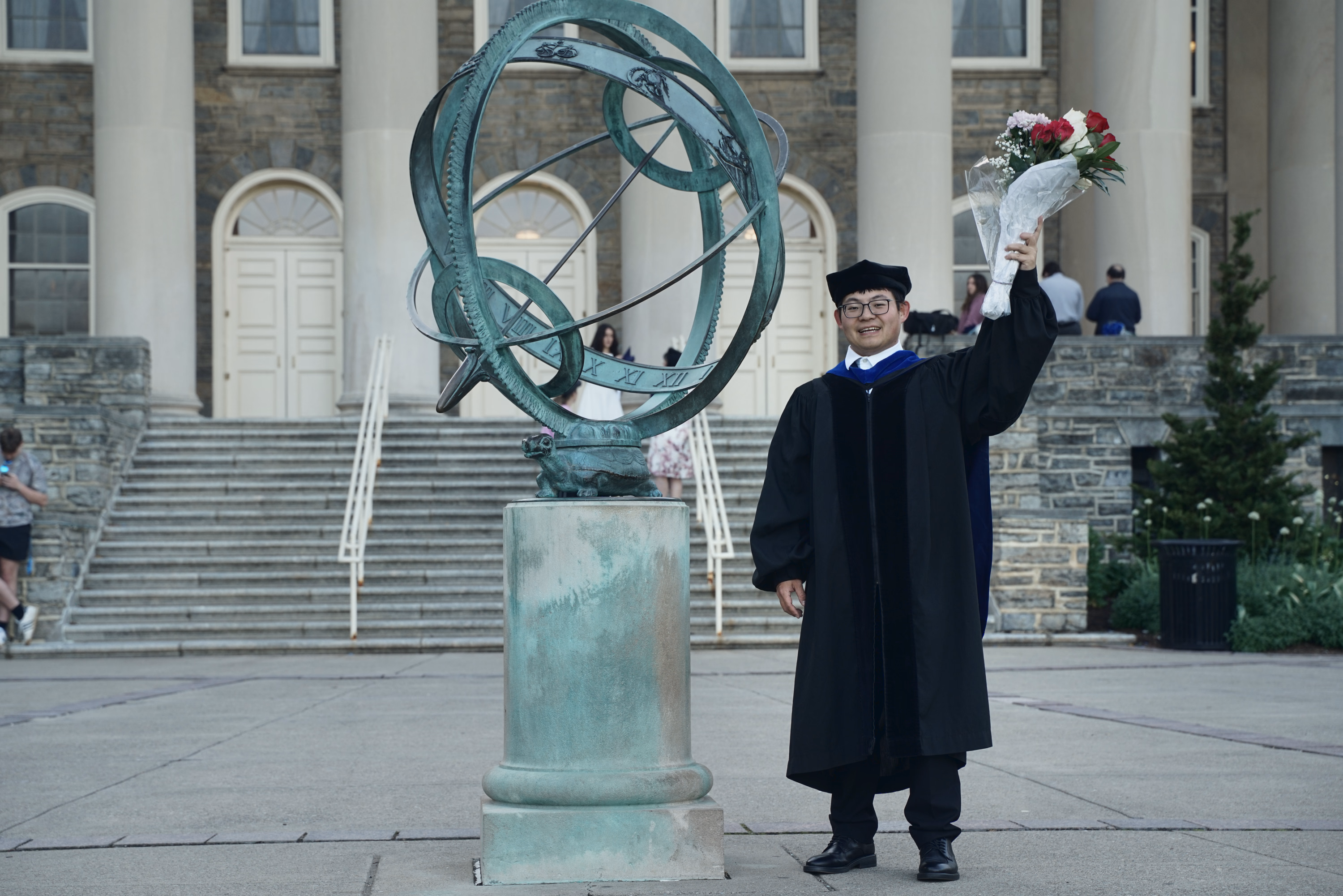 Graduate raising a bouquet beside the Old Main armillary sphere sculpture.