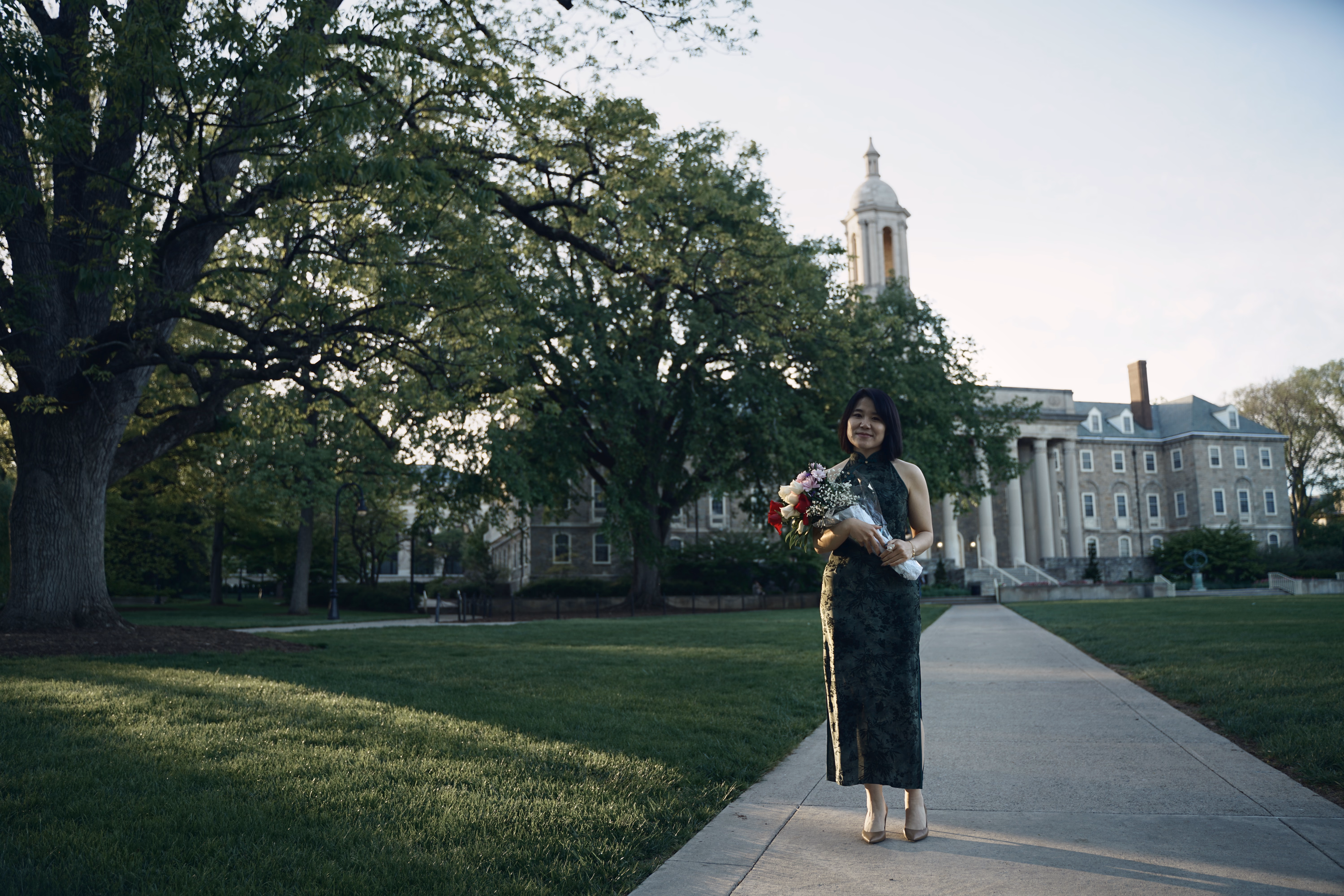 Woman in a dark green dress beneath a huge tree with Old Main tower across the lawn.