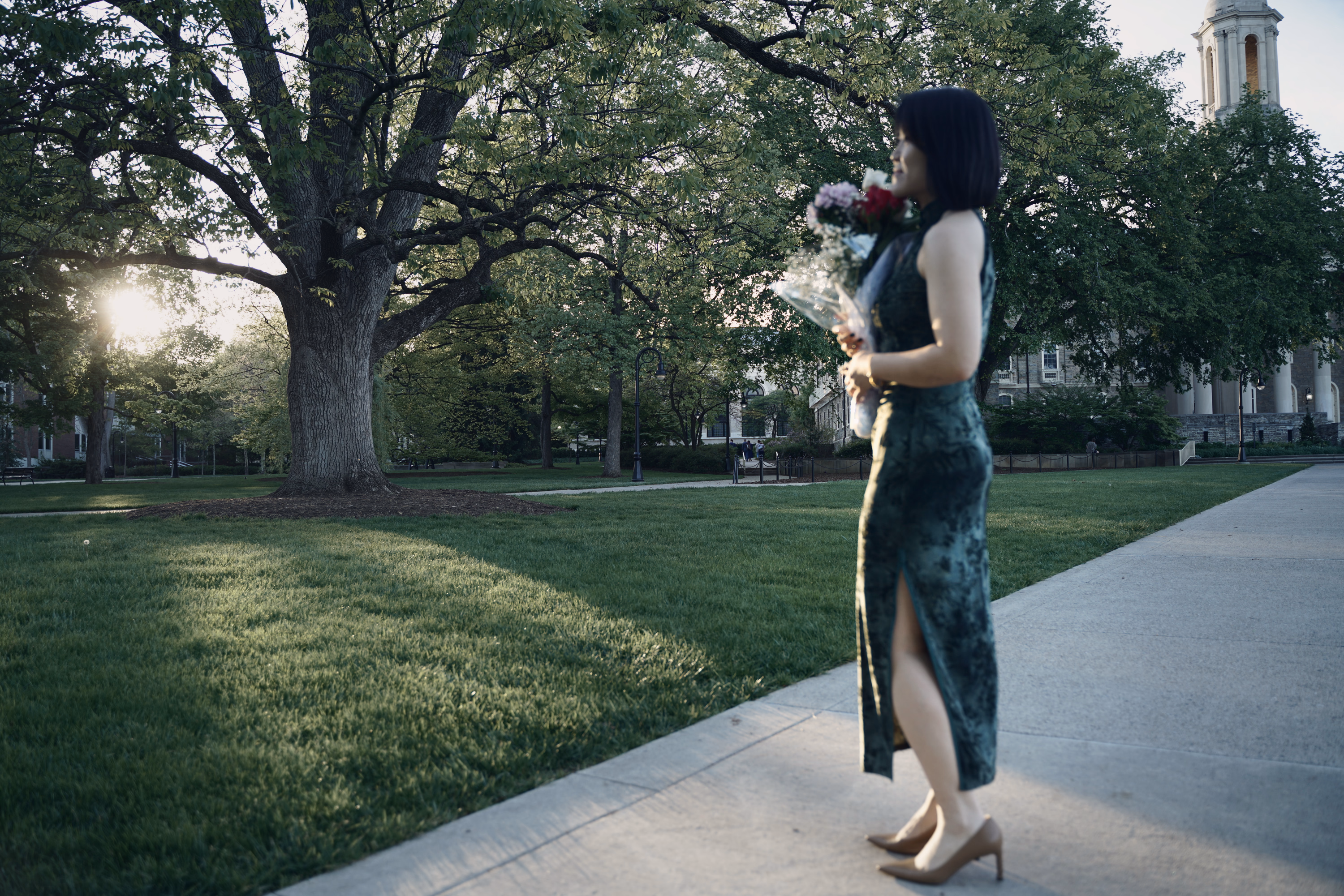 Soft profile of a woman holding flowers in front of a monumental tree and low sun near Old Main.