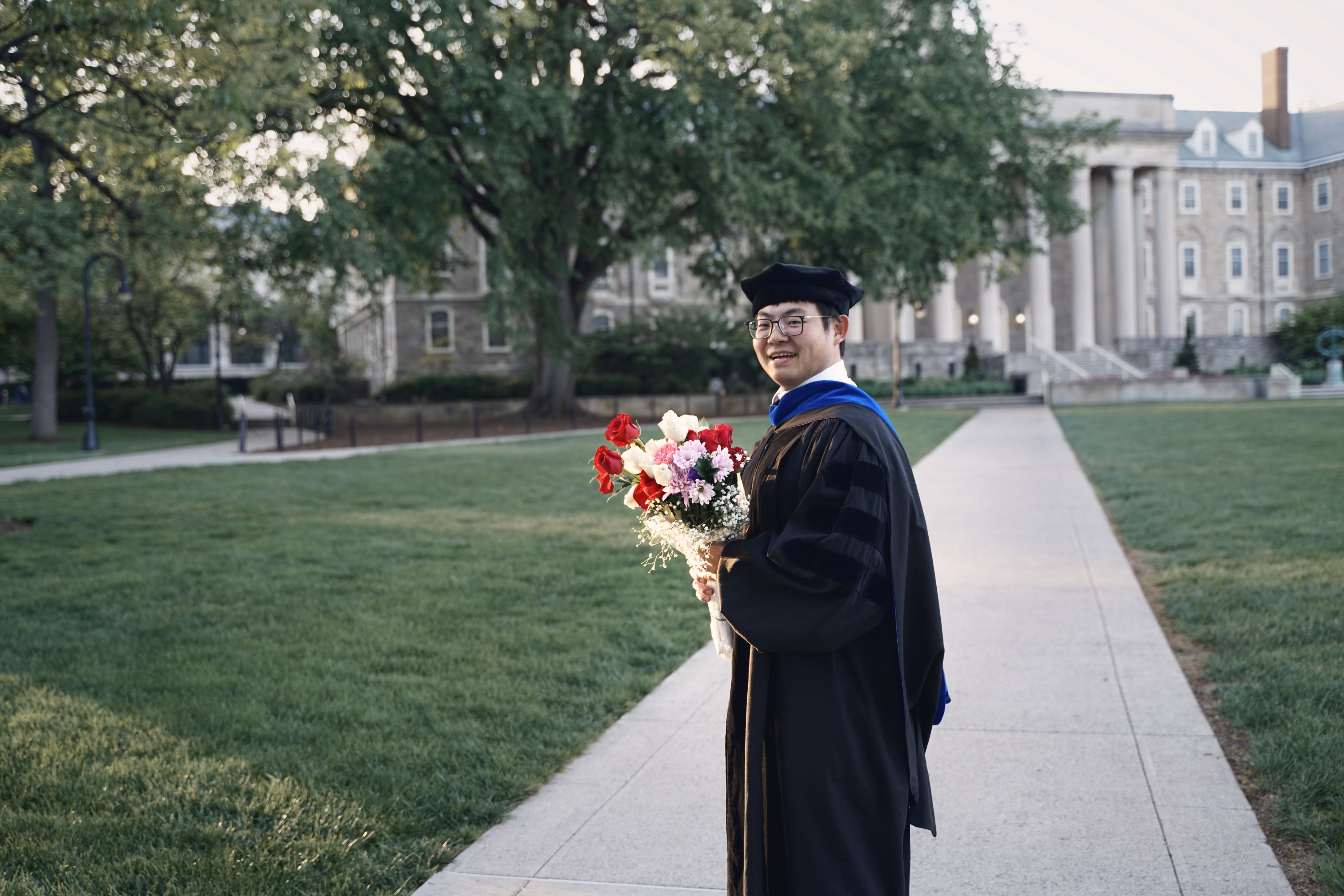 Graduate turning back toward the camera with a bouquet on a tree-lined campus path.