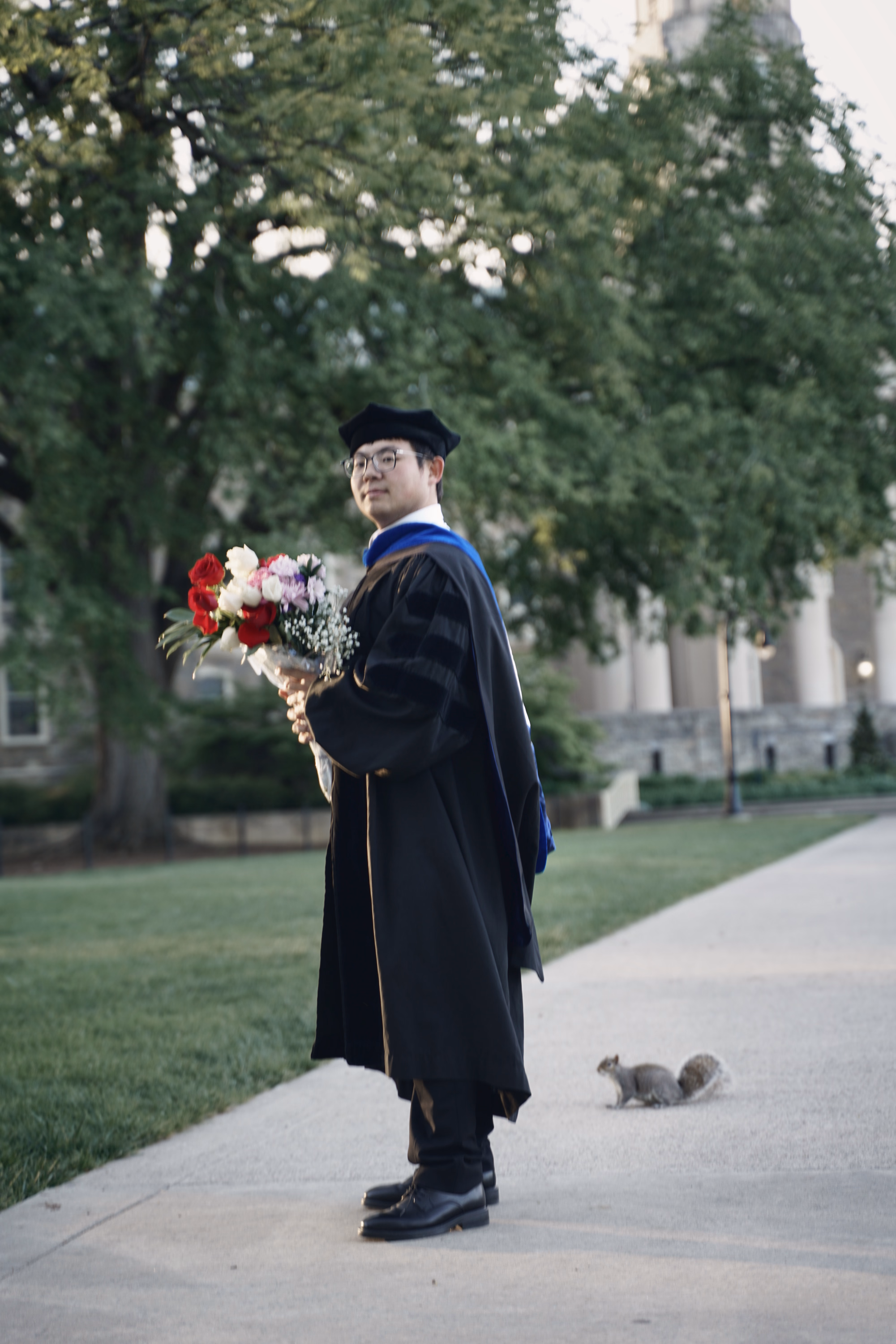 Graduate with flowers standing on a campus path as a squirrel sits nearby.