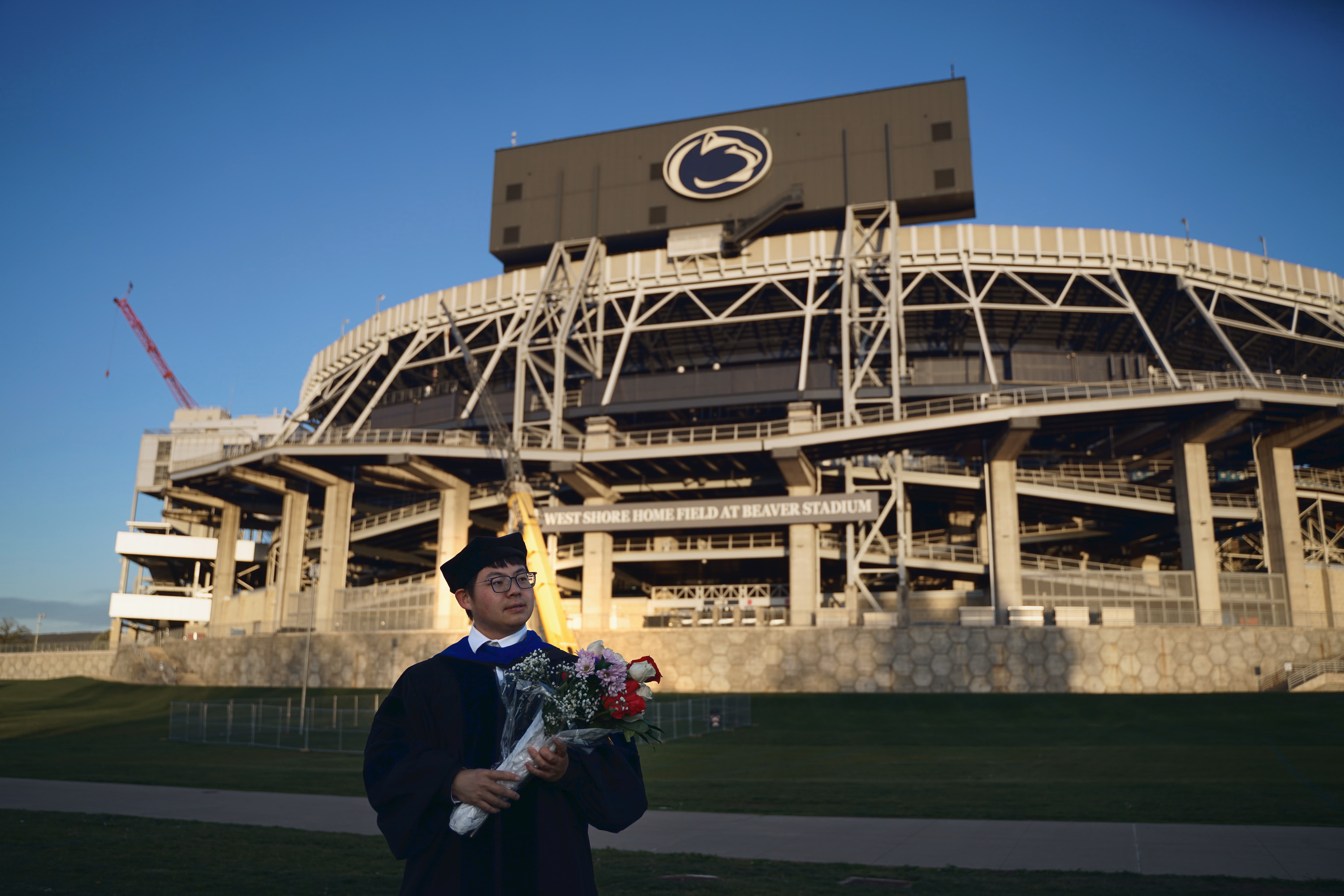 Wide environmental portrait of a graduate with flowers in front of Beaver Stadium.