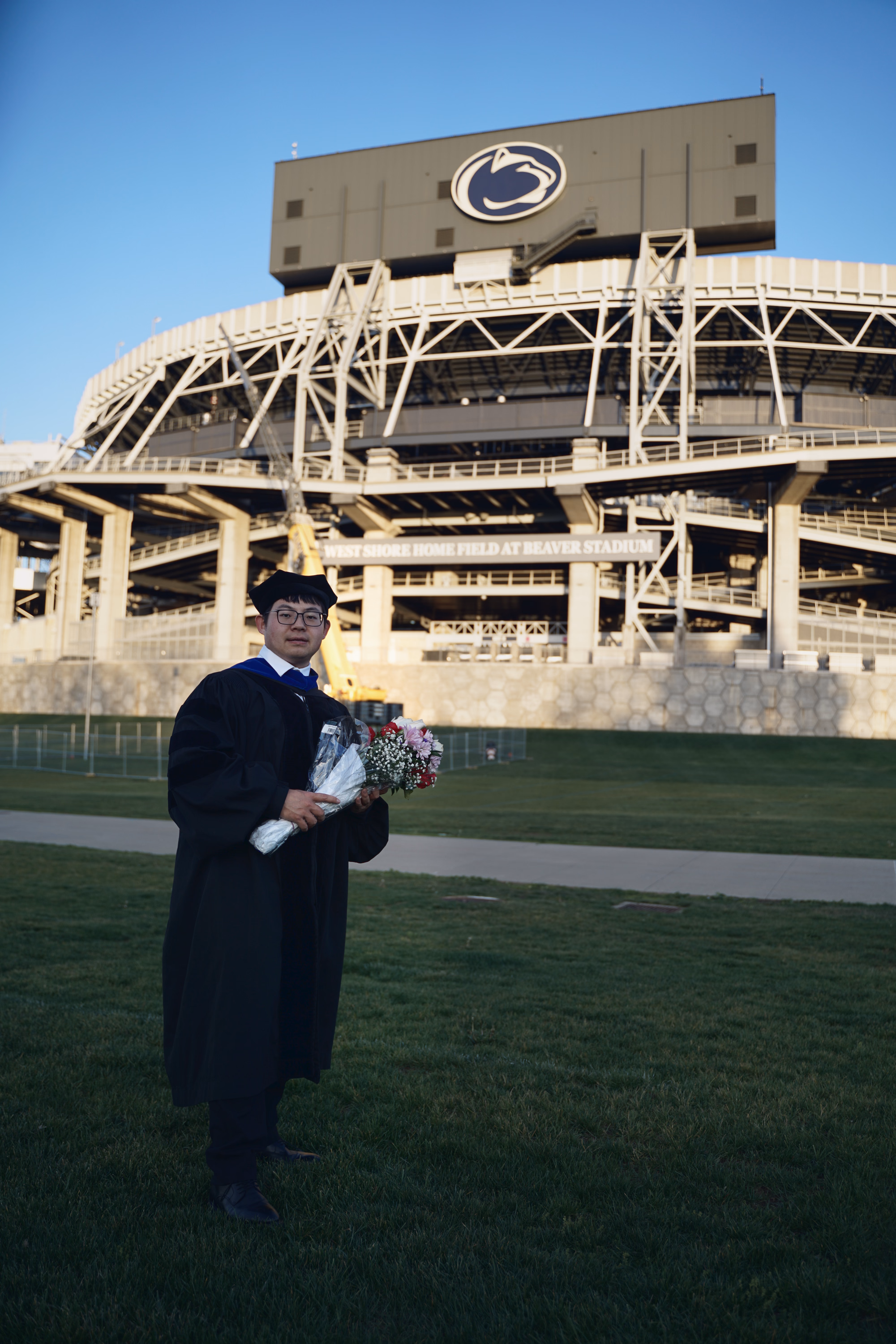Full-length graduate with bouquet standing in front of Beaver Stadium and the Nittany Lion logo.