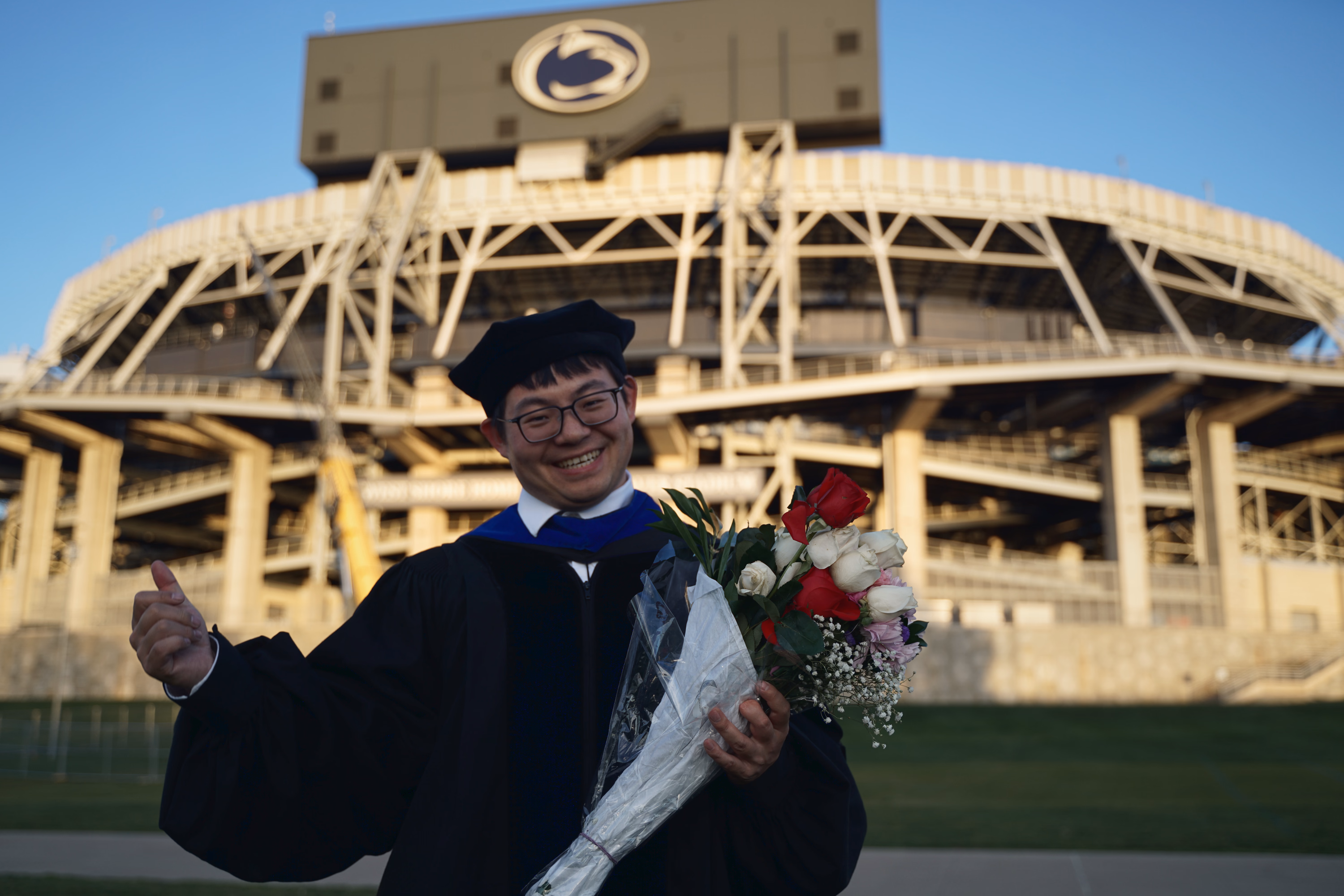 Joyful graduate holding flowers and giving a thumbs-up with Beaver Stadium softly behind.