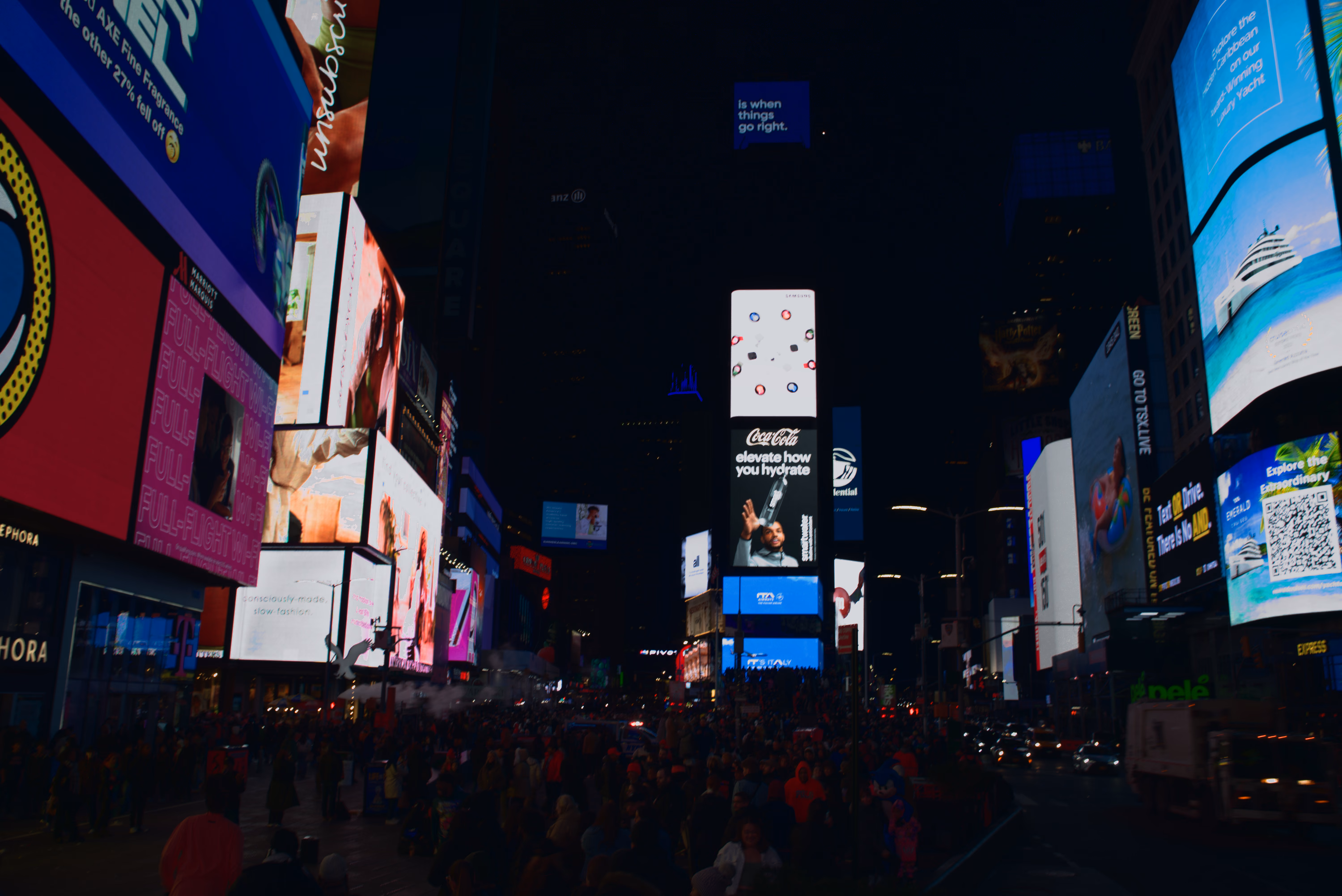Magenta and cyan Times Square screens hang above moving traffic and a packed pedestrian plaza.