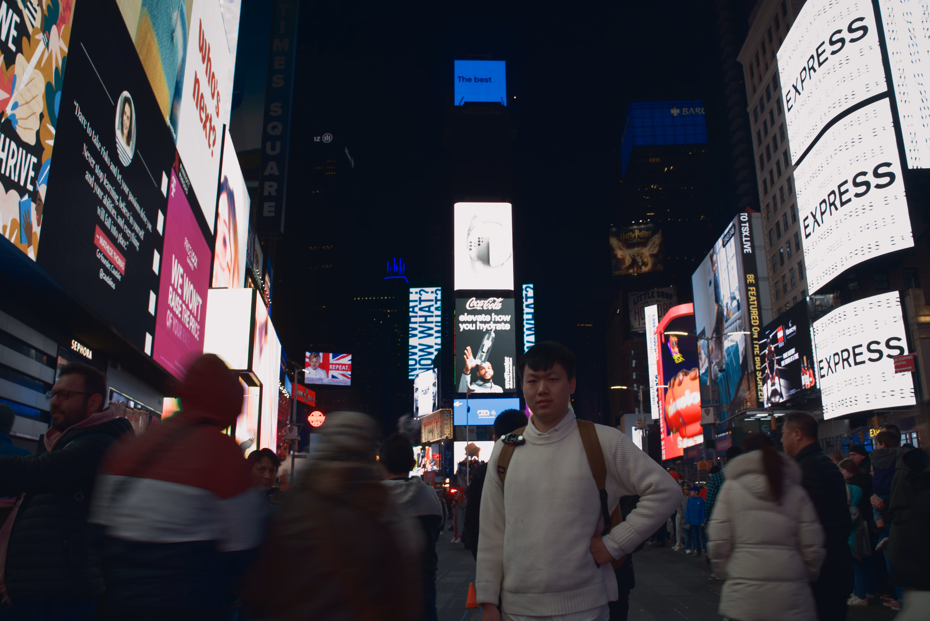 A man in a white sweater stands still in Times Square while pedestrians blur around him.