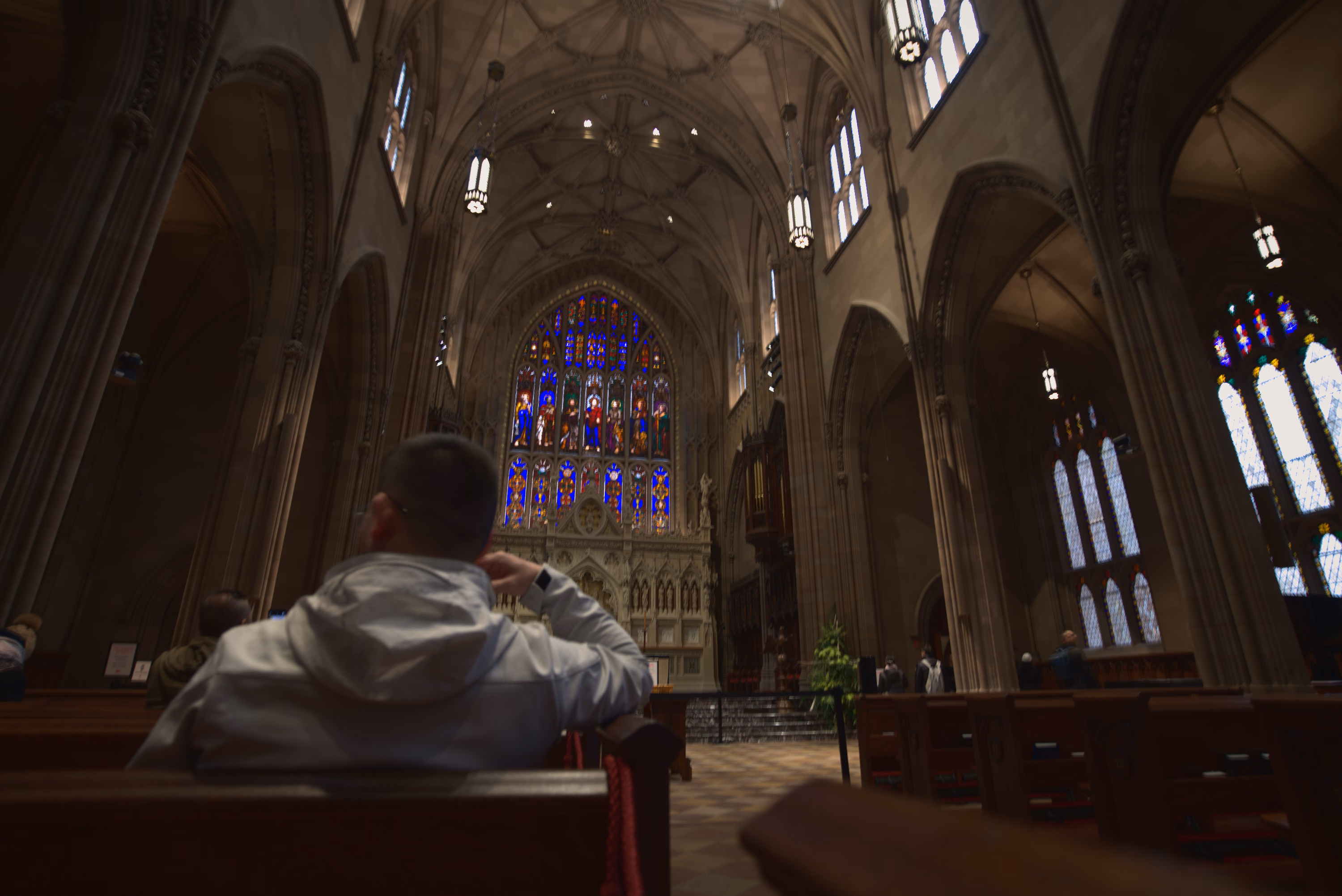 A seated visitor looks toward a stained-glass altar beneath the high stone vaults of a New York church.