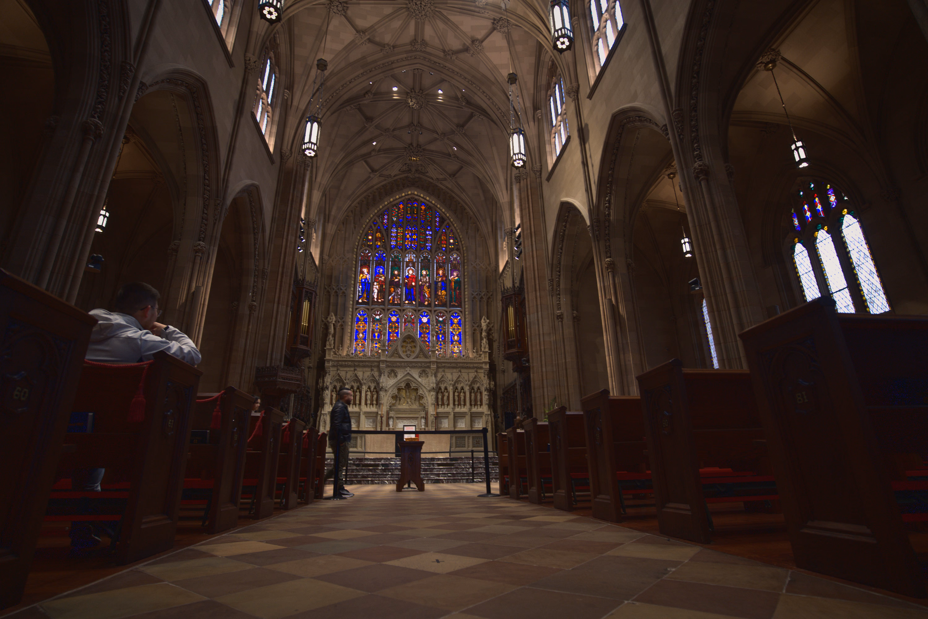 A low central aisle leads through dark wooden pews toward a jewel-colored stained-glass altar.