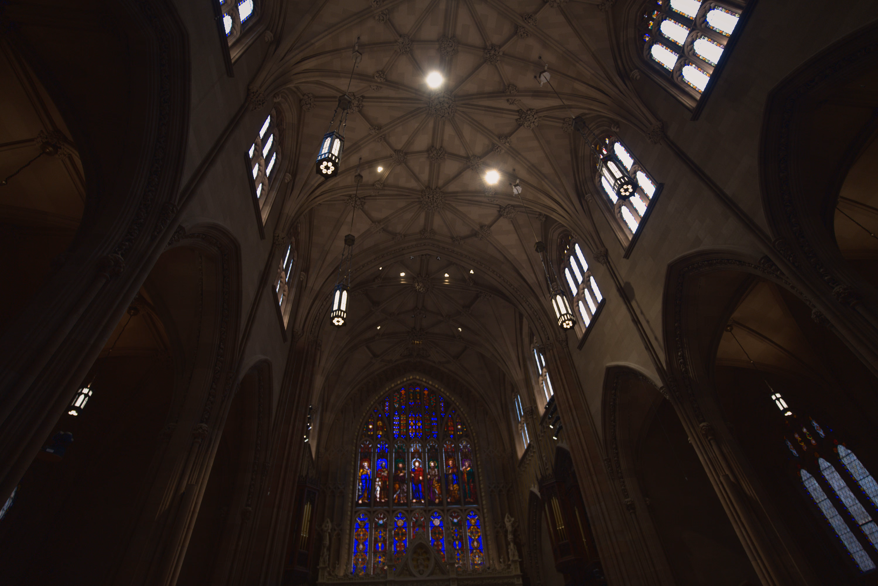 Stone ribs, hanging lanterns, and bright windows converge above a band of stained glass.