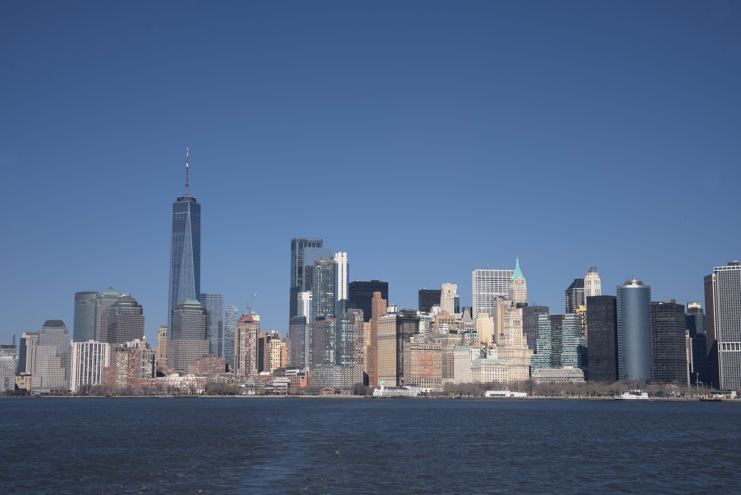 Lower Manhattan sits low beneath an expansive winter-blue sky and a narrow band of harbor water.