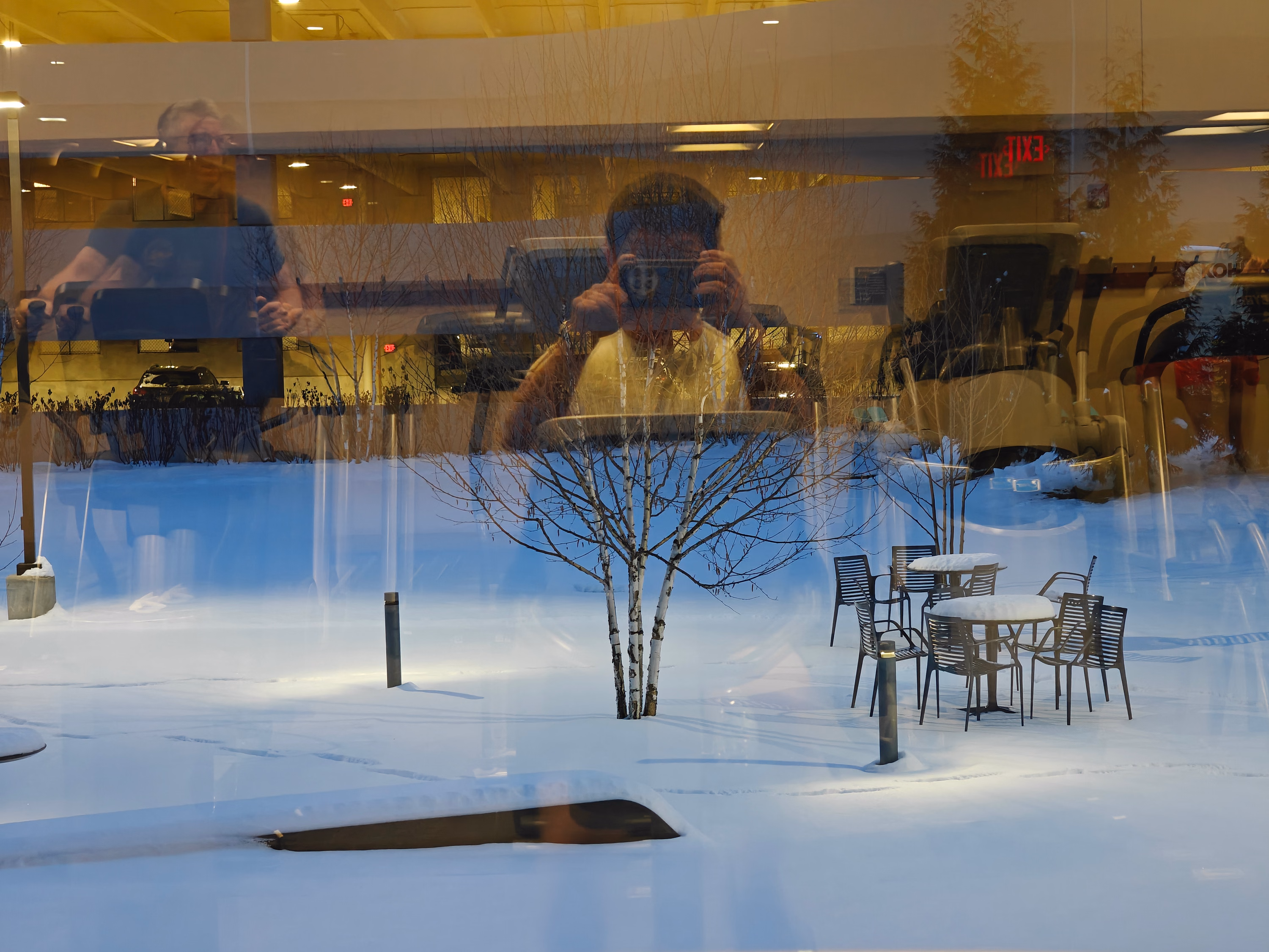 A photographer and warm interior lights overlap a snowy courtyard in a window reflection.