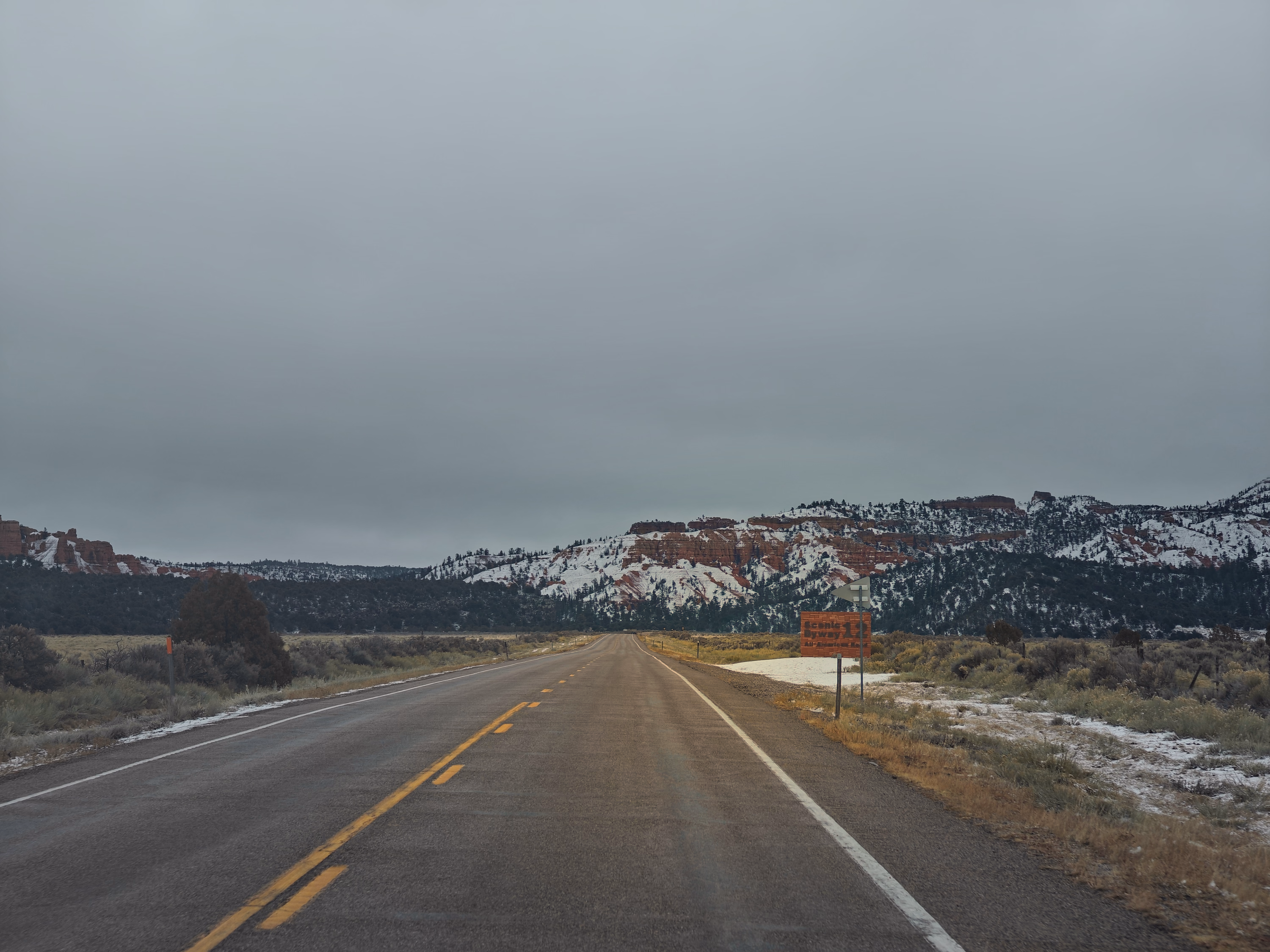 An empty highway points toward snow-striped red mesas beneath a vast gray winter sky.