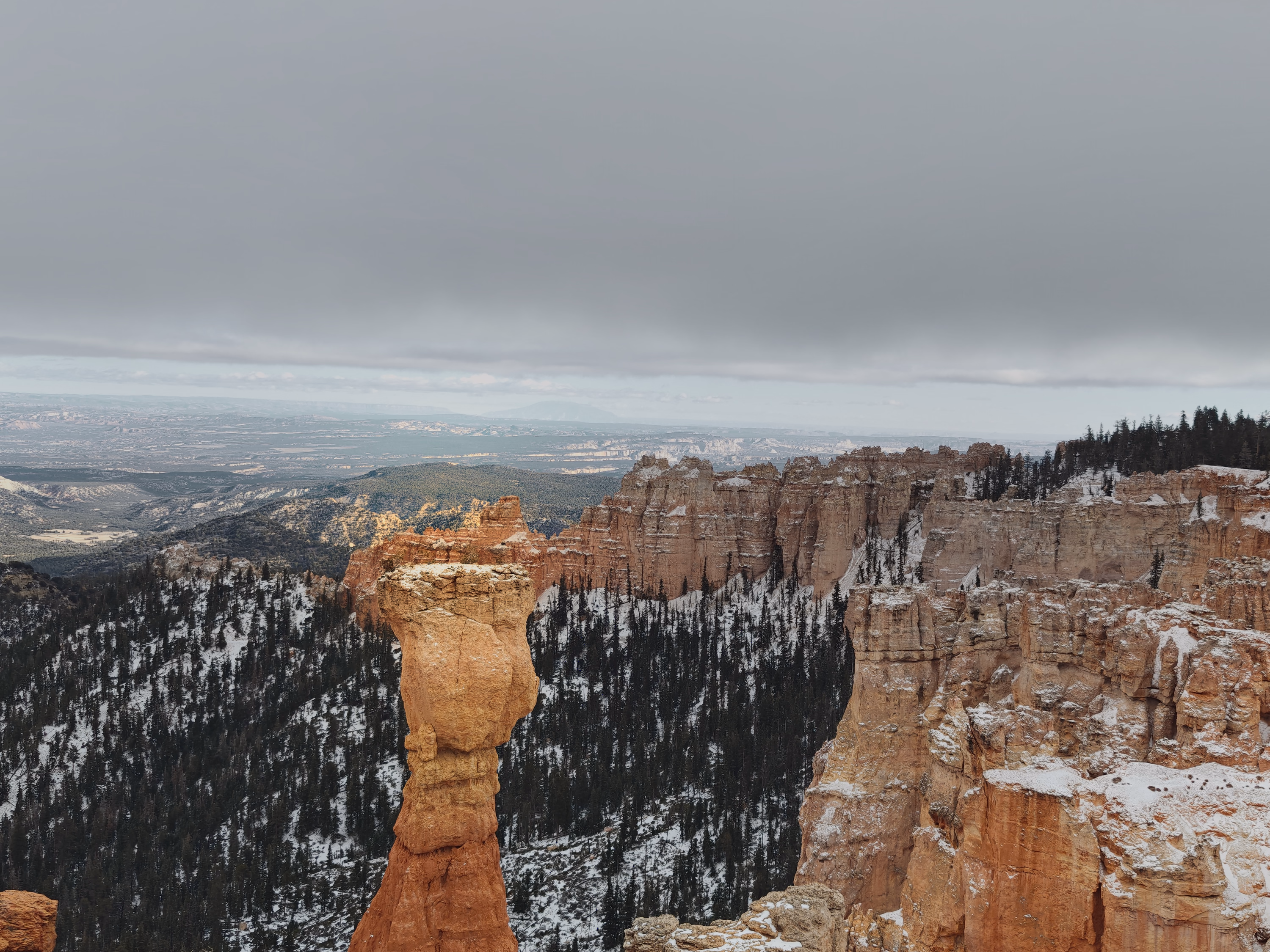 A solitary hoodoo rises against gray sky above snowy walls and a distant pale desert.