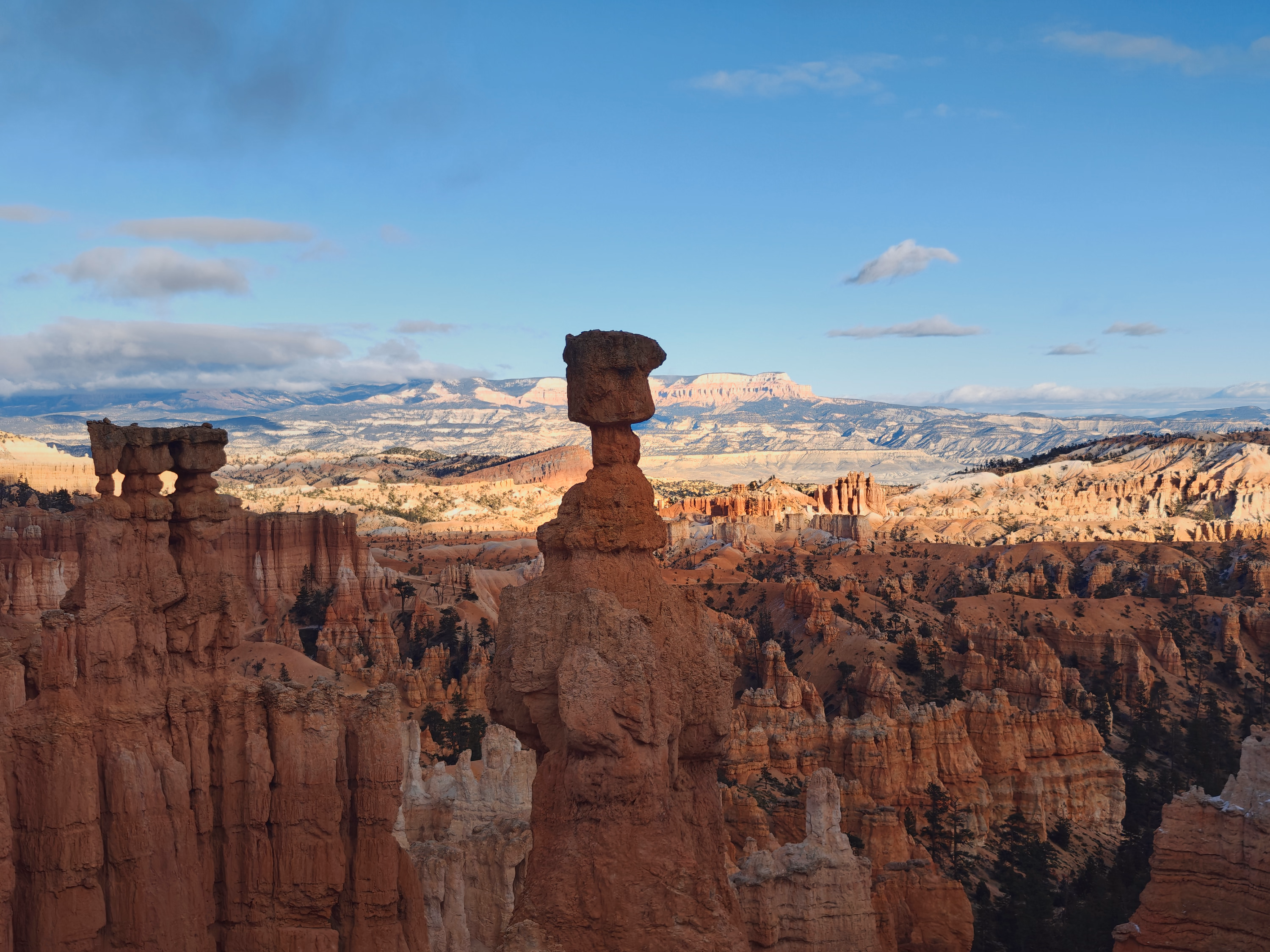 A mushroom-shaped hoodoo stands against layered red basins and a snow-bright horizon.