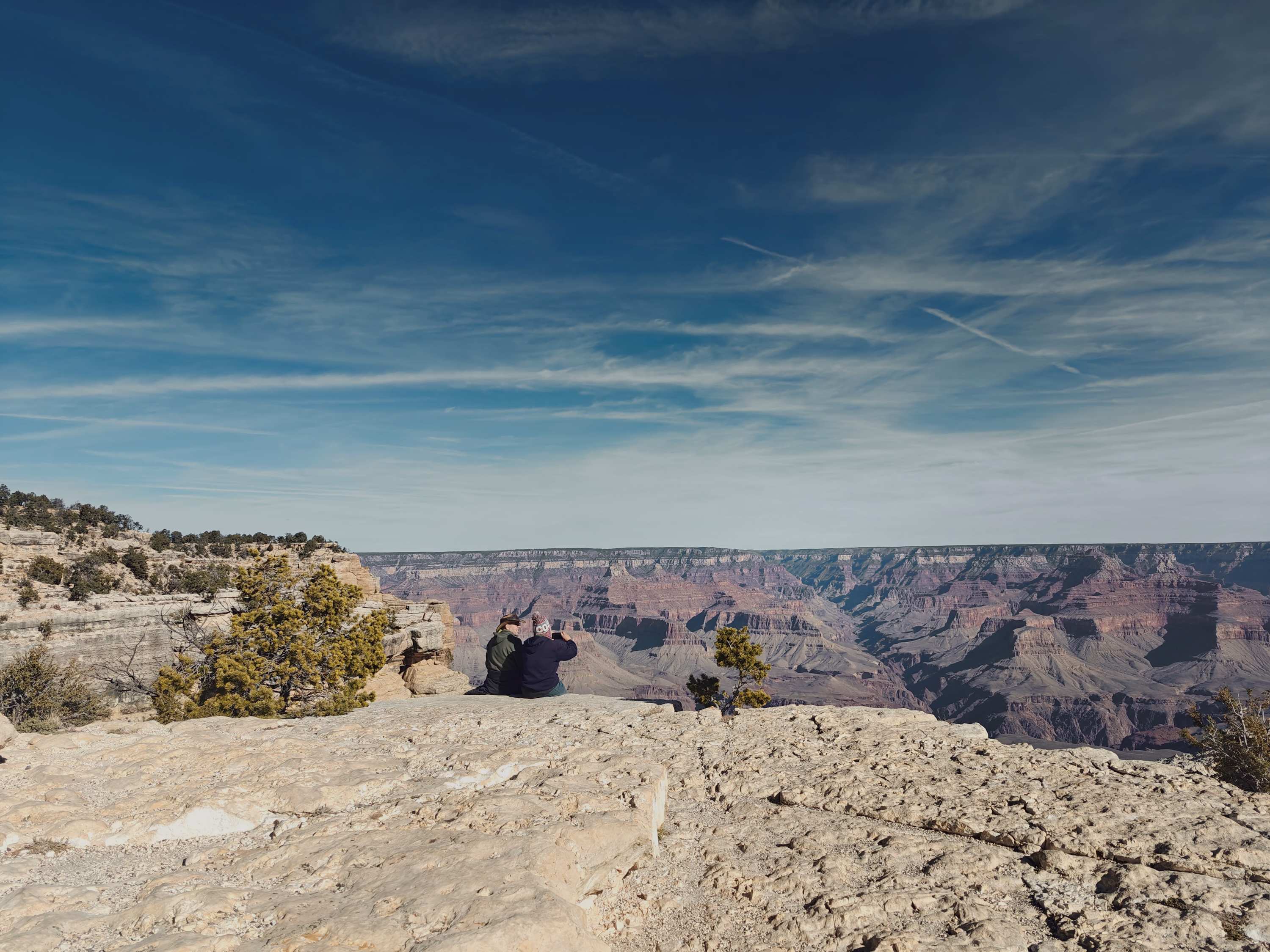 Two visitors sit at the Grand Canyon rim, small against rock, distance, and a muted blue sky.