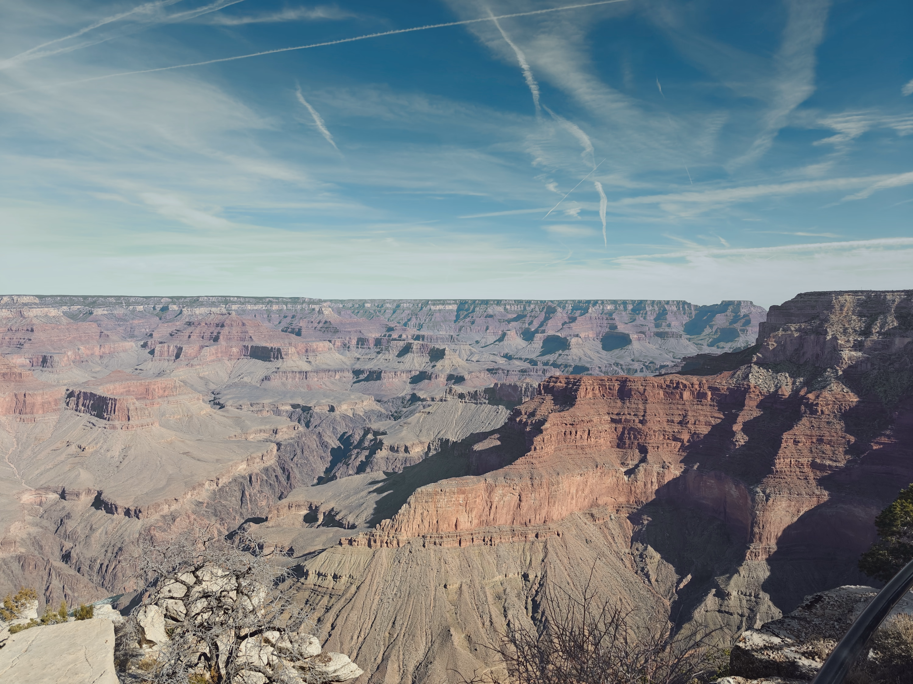 Contrails cross a muted blue sky above terraced Grand Canyon walls and deep side canyons.