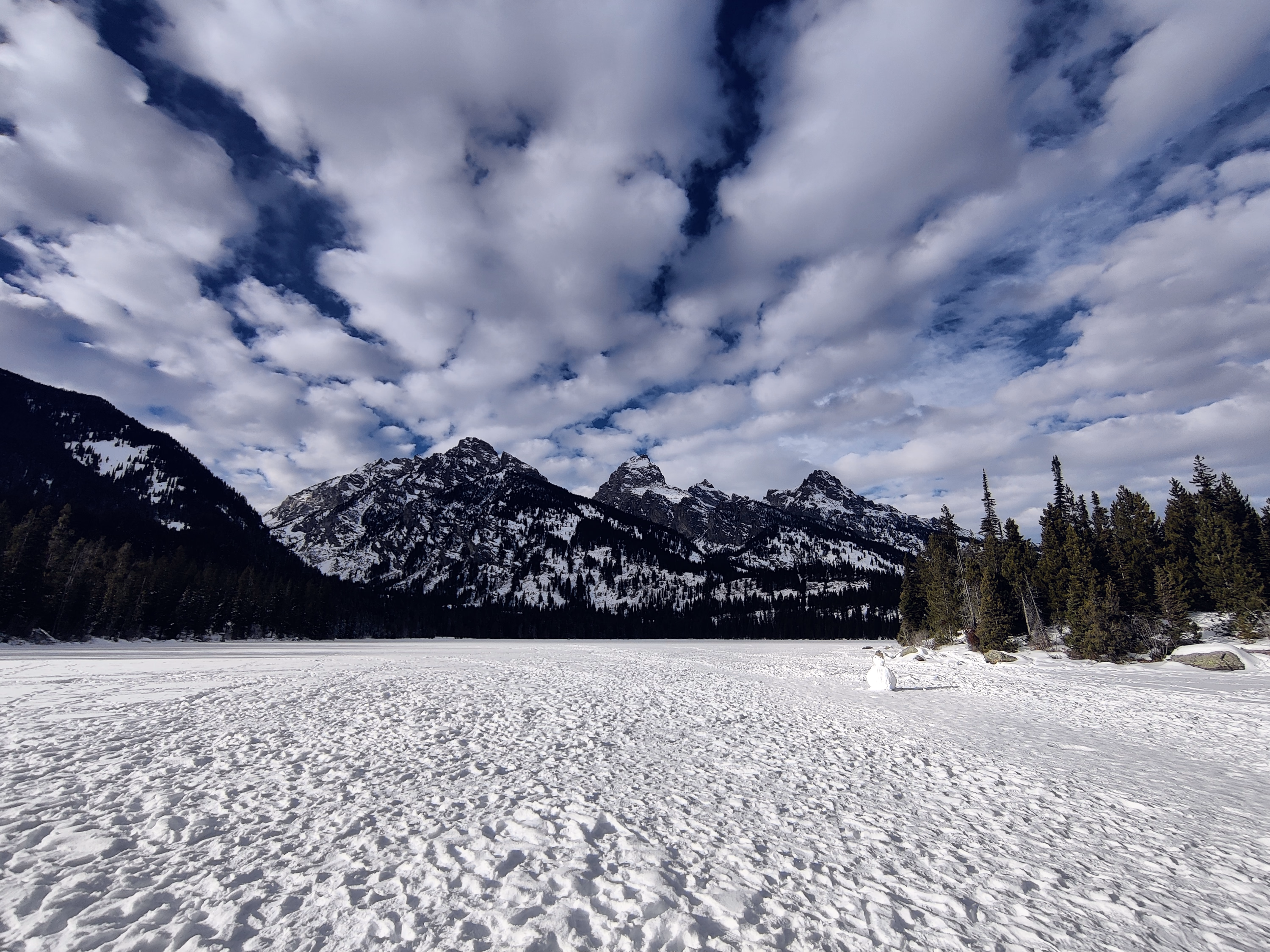 A tiny snowman stands at the edge of a frozen lake beneath mountains and a vast cloud field.