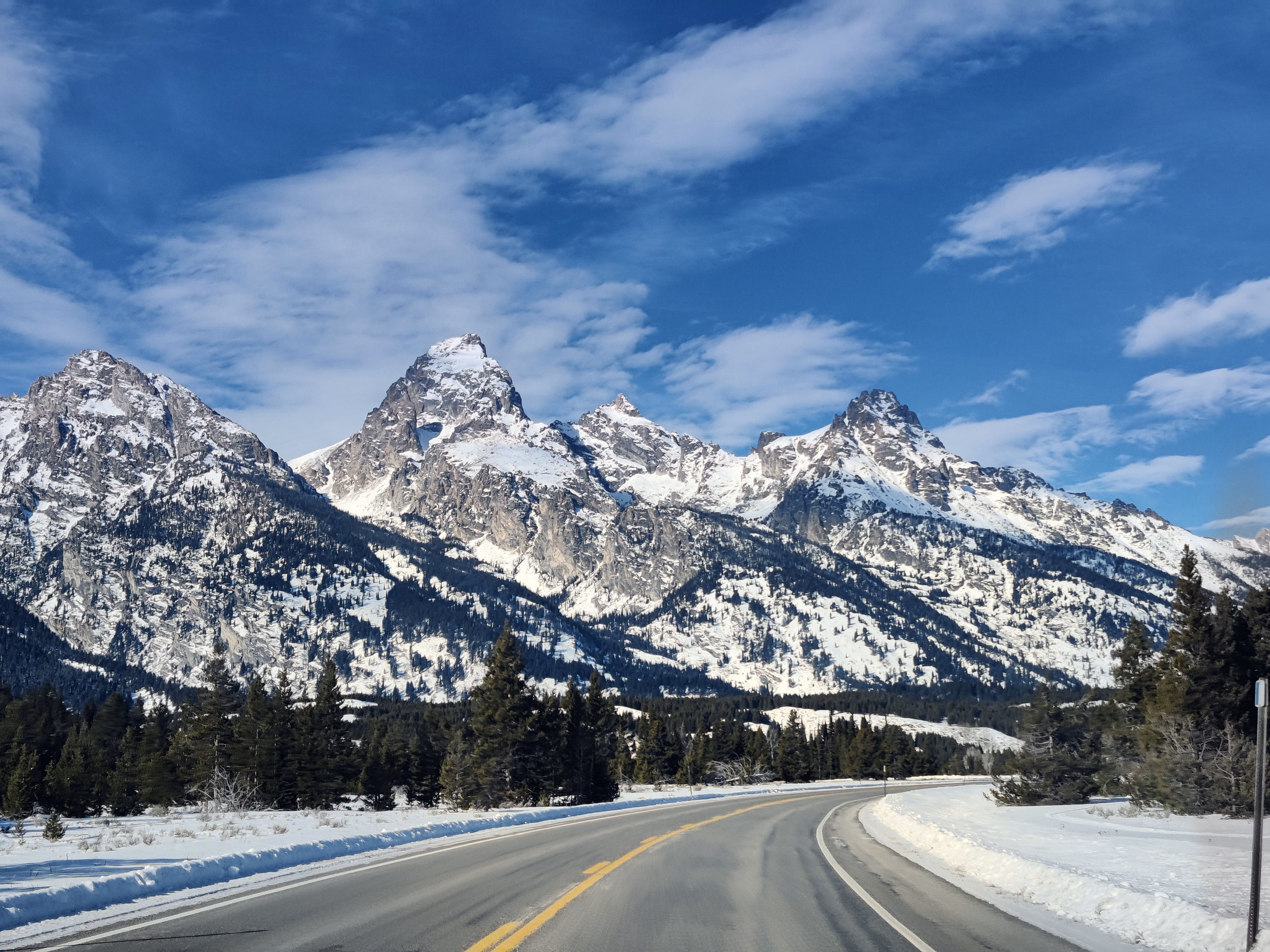 A winter road crosses open snow toward the jagged Grand Teton skyline.