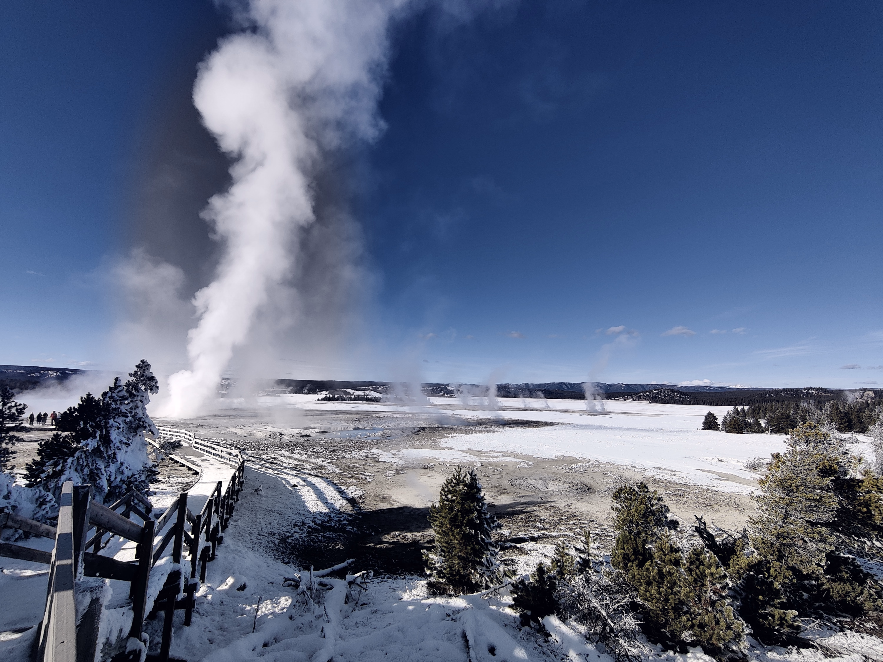 Visitors and a boardwalk descend toward a towering steam vent across the frozen basin.