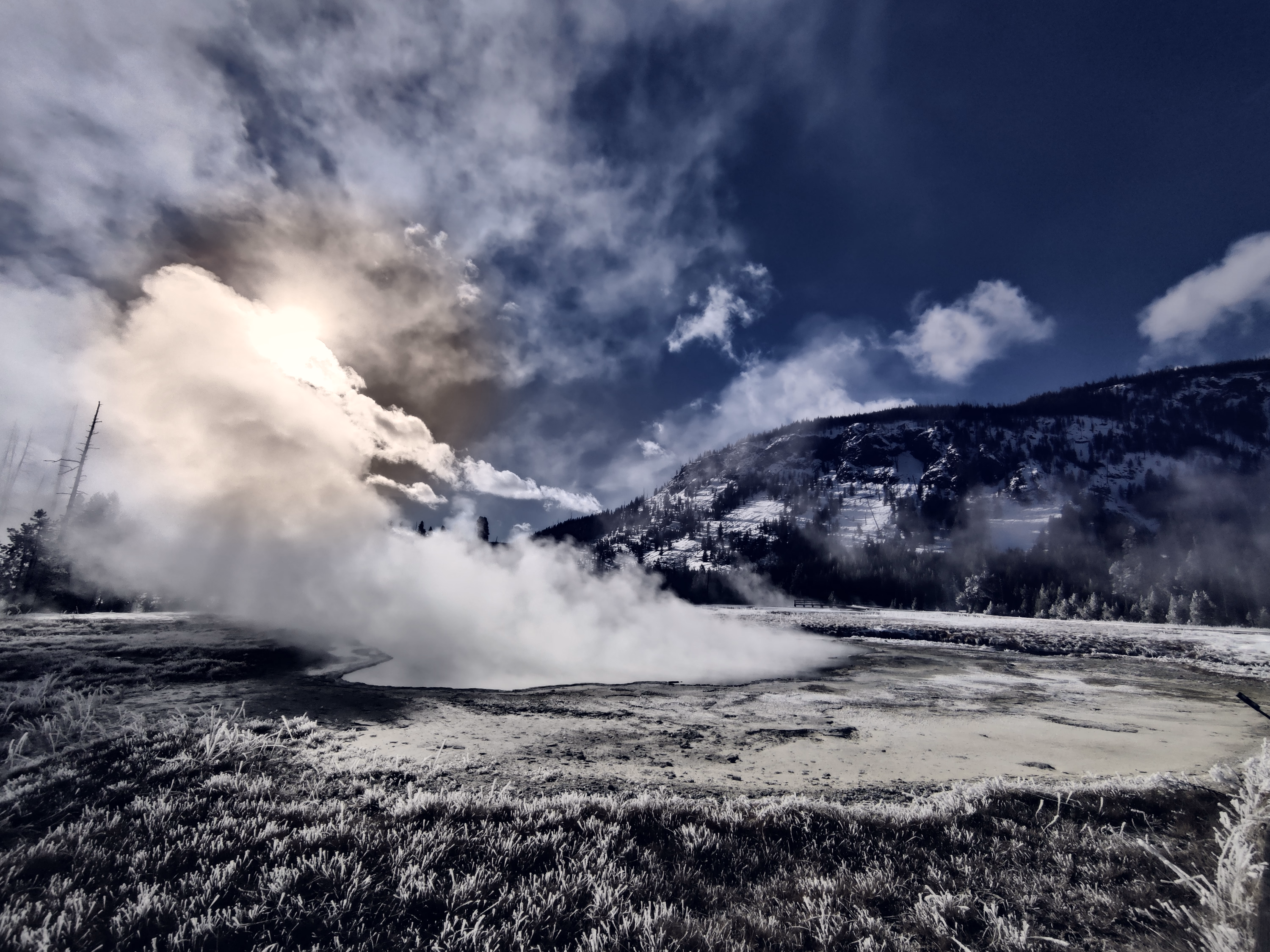 Backlit steam pours across a geothermal pool below a dark snowy ridge and fractured sky.