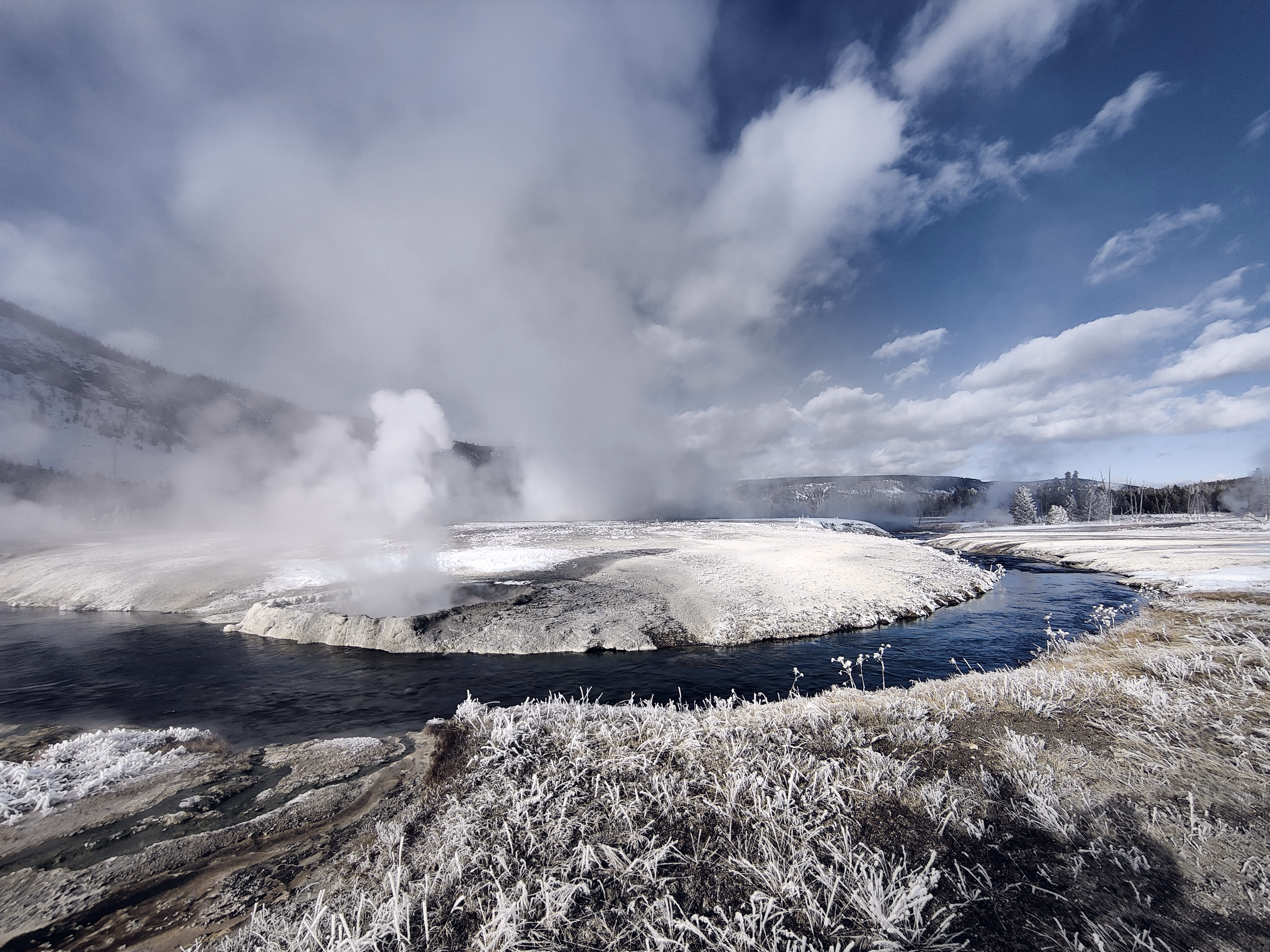 A dark river bends around a frost-white geothermal island beneath rising steam and cloud.