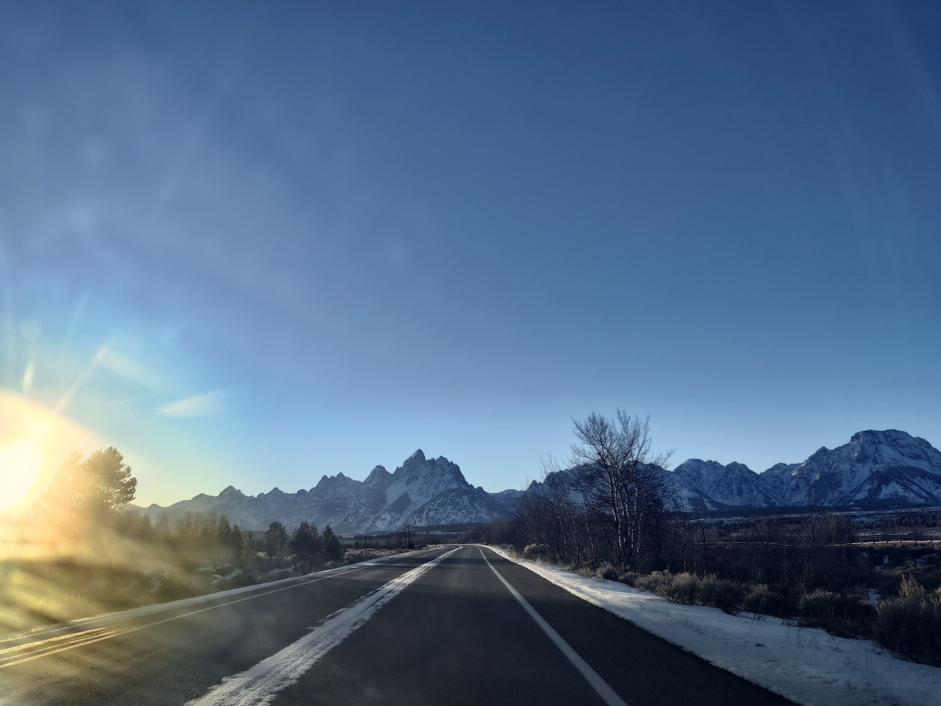 A road carries the viewer back toward the Tetons through low winter sun and windshield haze.